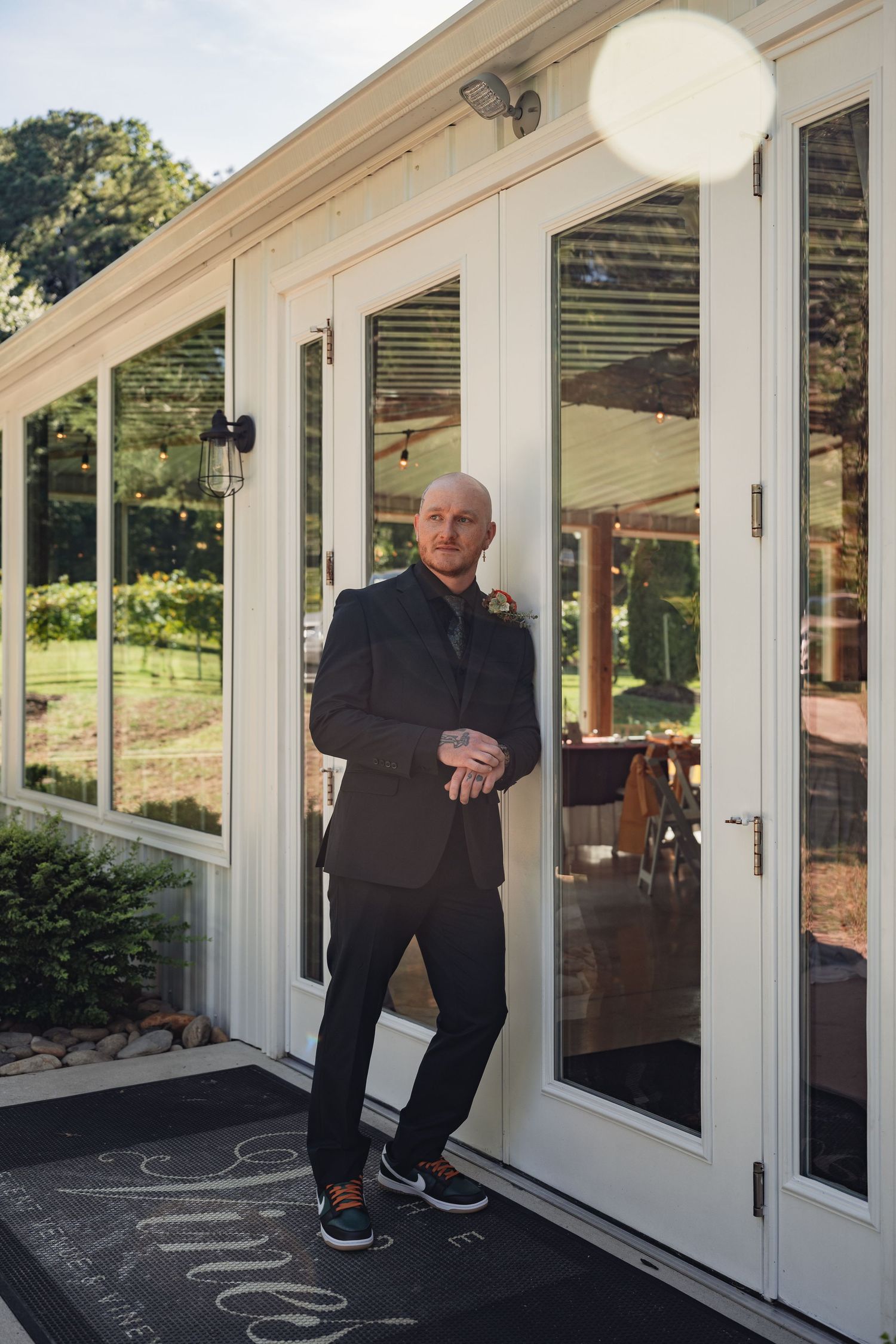 Bald groom in black suit leaning against glass venue doors on wedding day.