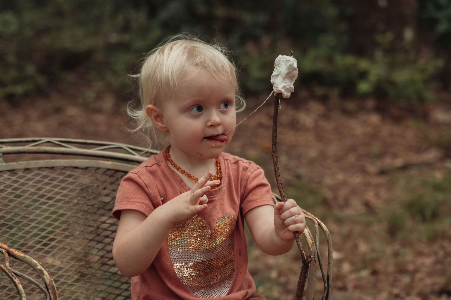 Toddler girl with blonde hair holding a roasted marshmallow on a stick outdoors, wearing a pink shirt.