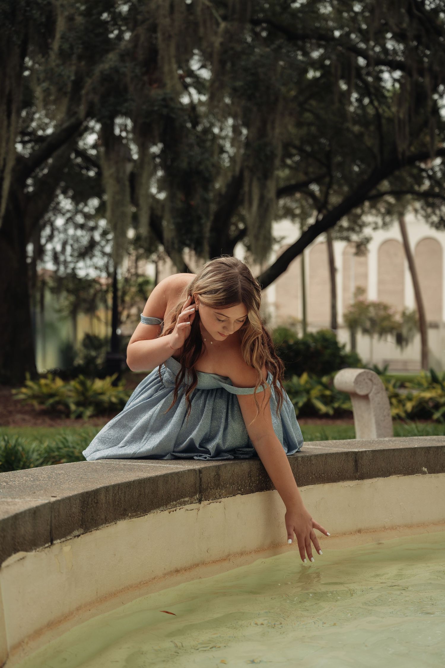 Young woman in blue off-shoulder dress touching fountain water in a lush garden with Spanish moss trees.