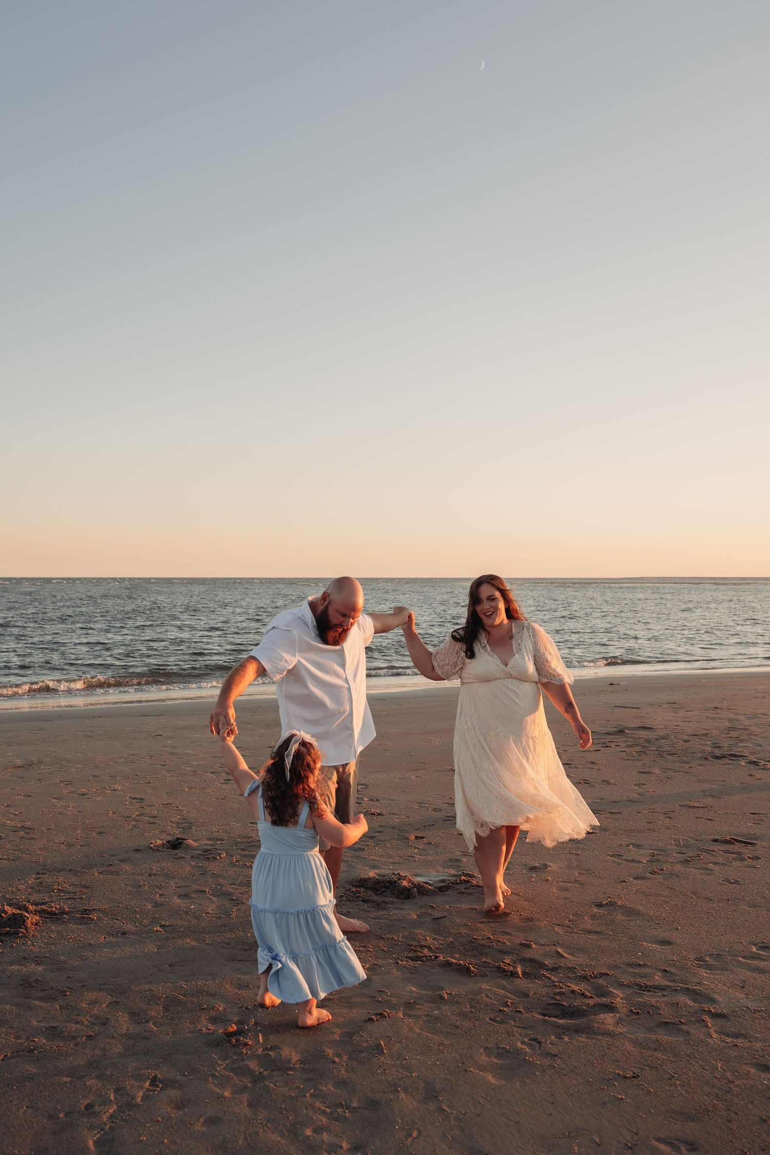 Family of three holding hands and dancing on a sandy beach at sunset, with a woman in a cream dress, a man and a young girl.