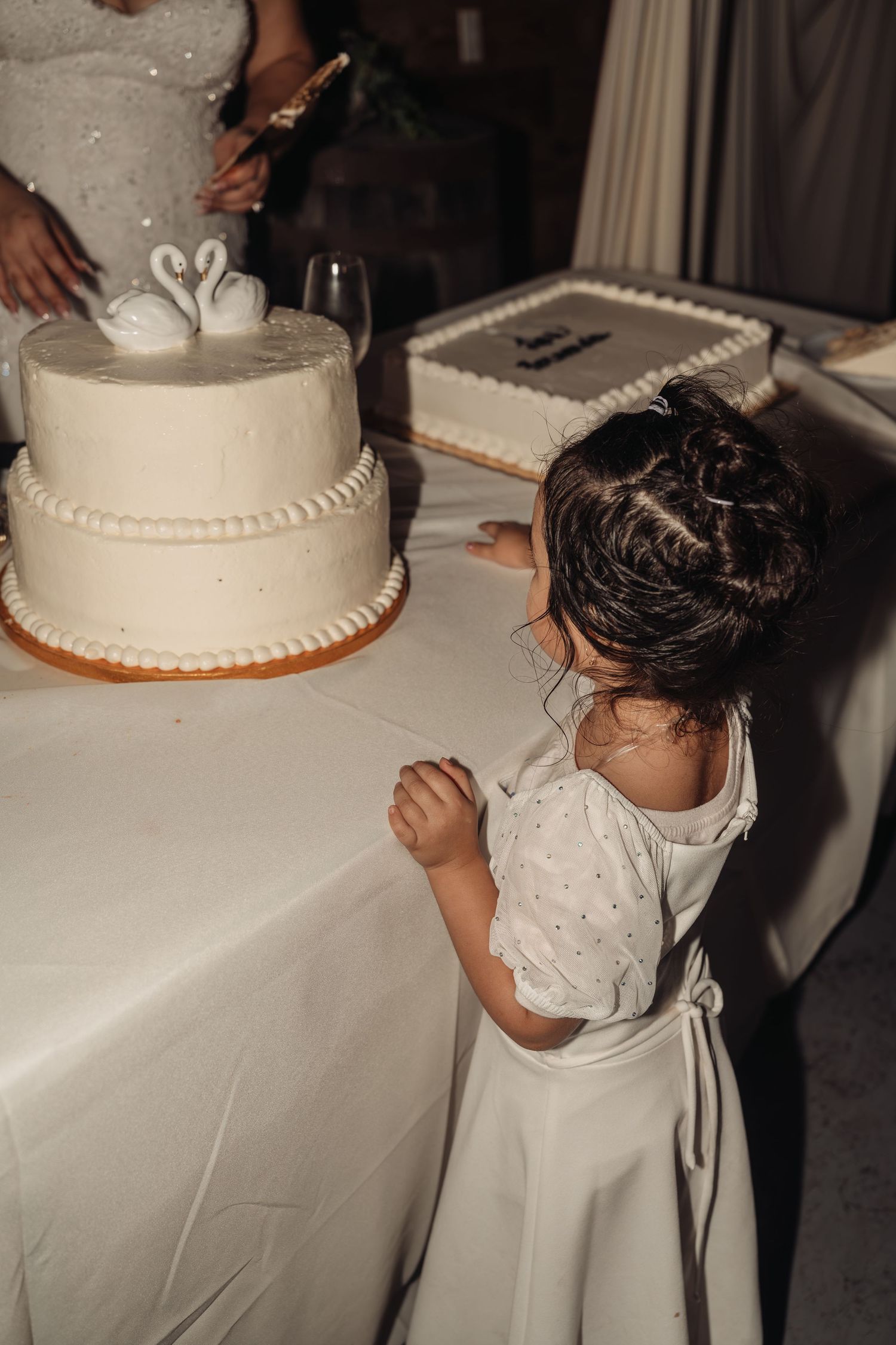 A young flower girl in a white dress reaches toward a two-tier wedding cake decorated with swan toppers at a reception.