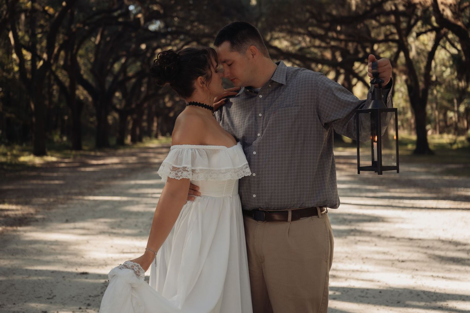 Couple sharing a tender kiss outdoors, woman in flowing white off-shoulder dress, man holding a black lantern in sunlit woods.