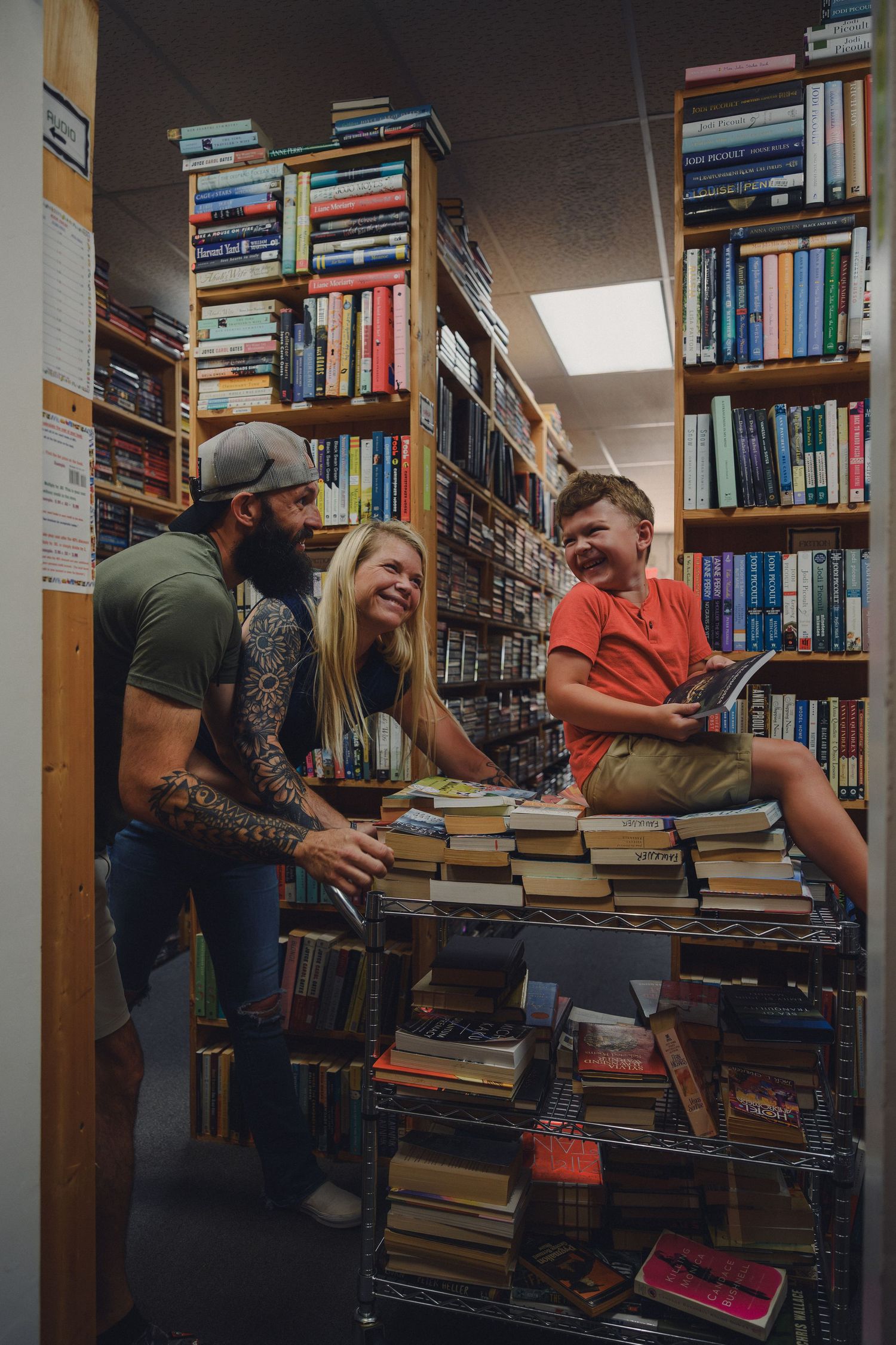 Family browsing books together in a cozy used bookstore, smiling among shelves and stacks of colorful books.