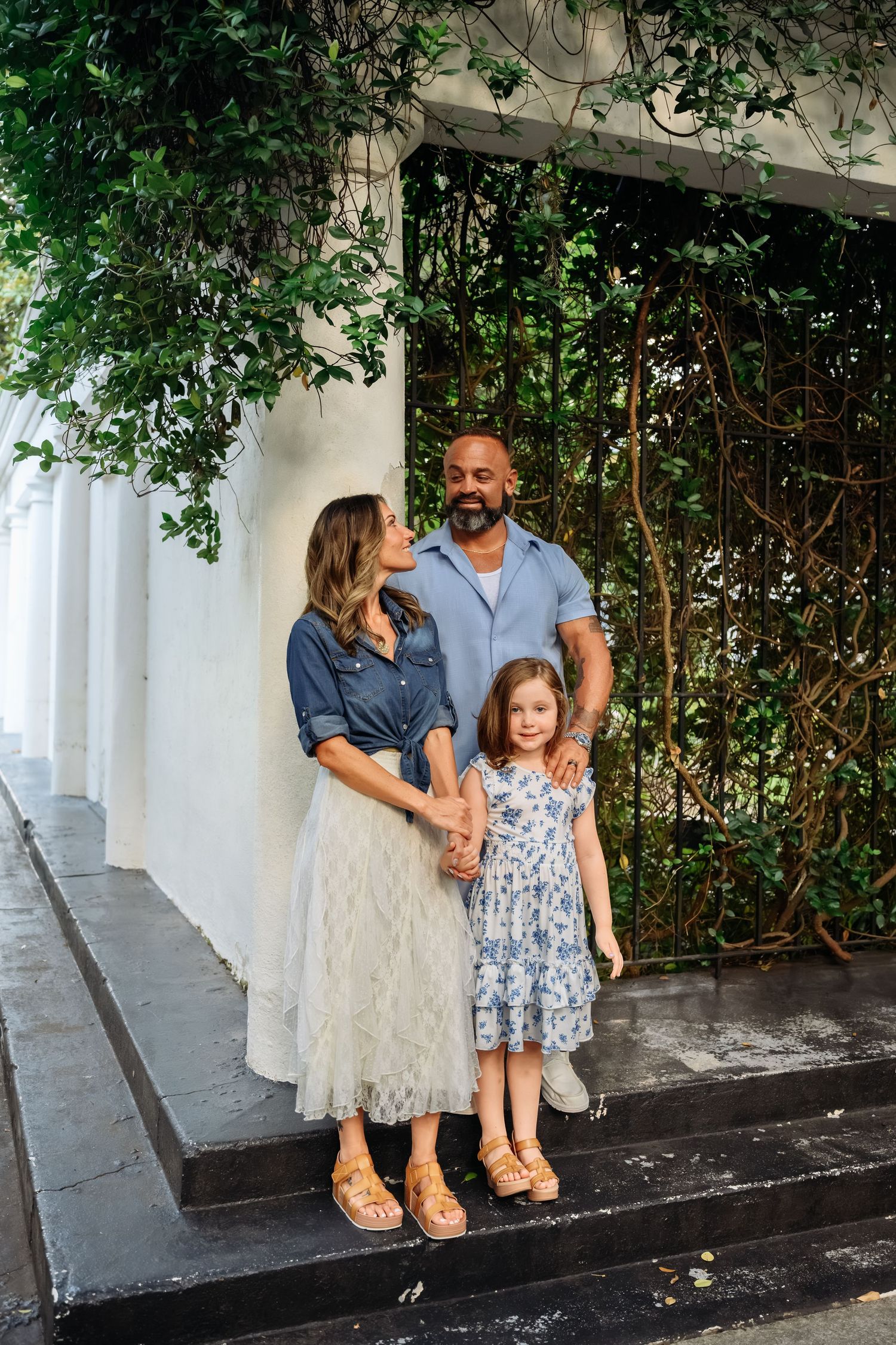A family of three poses on steps surrounded by lush greenery, wearing coordinating blue outfits.