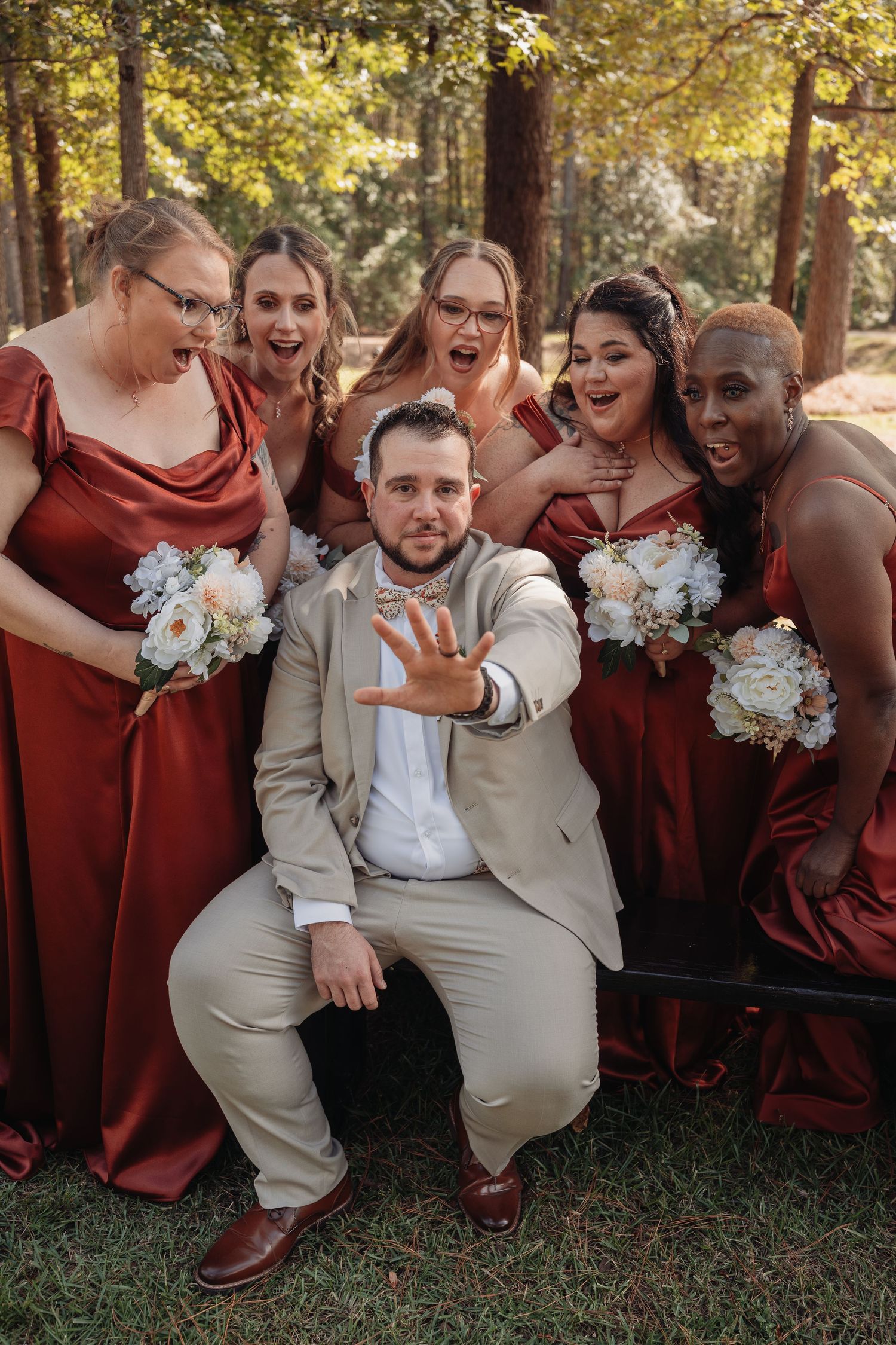 Groom in a tan suit crouches showing his ring as excited bridesmaids in rust red dresses holding white bouquets laugh behind him outdoors.