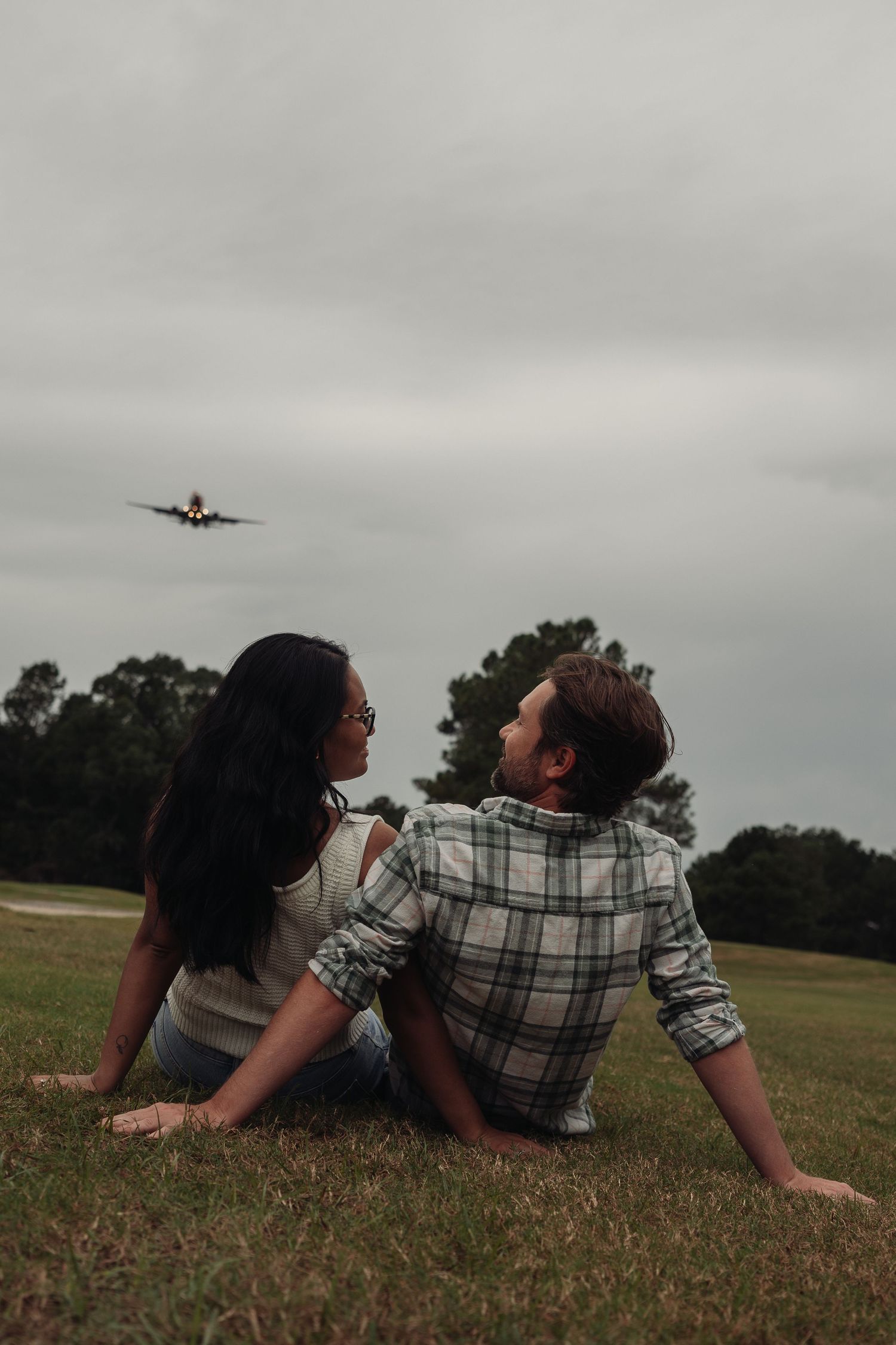 Couple sitting on grass field looking at each other with airplane flying overhead on a cloudy day.