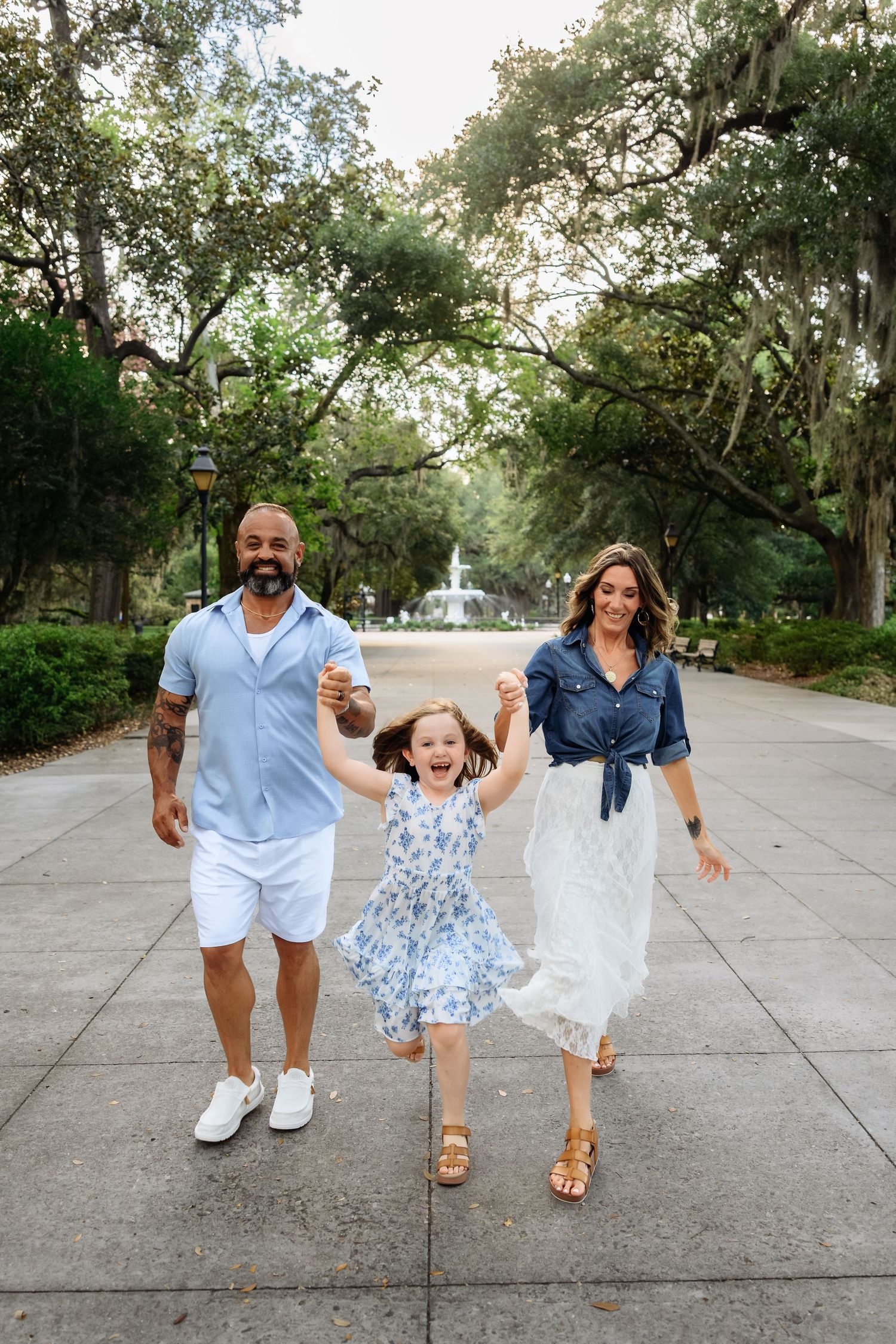 A joyful family of three walks hand-in-hand through a lush park path, with a fountain and trees in the background.