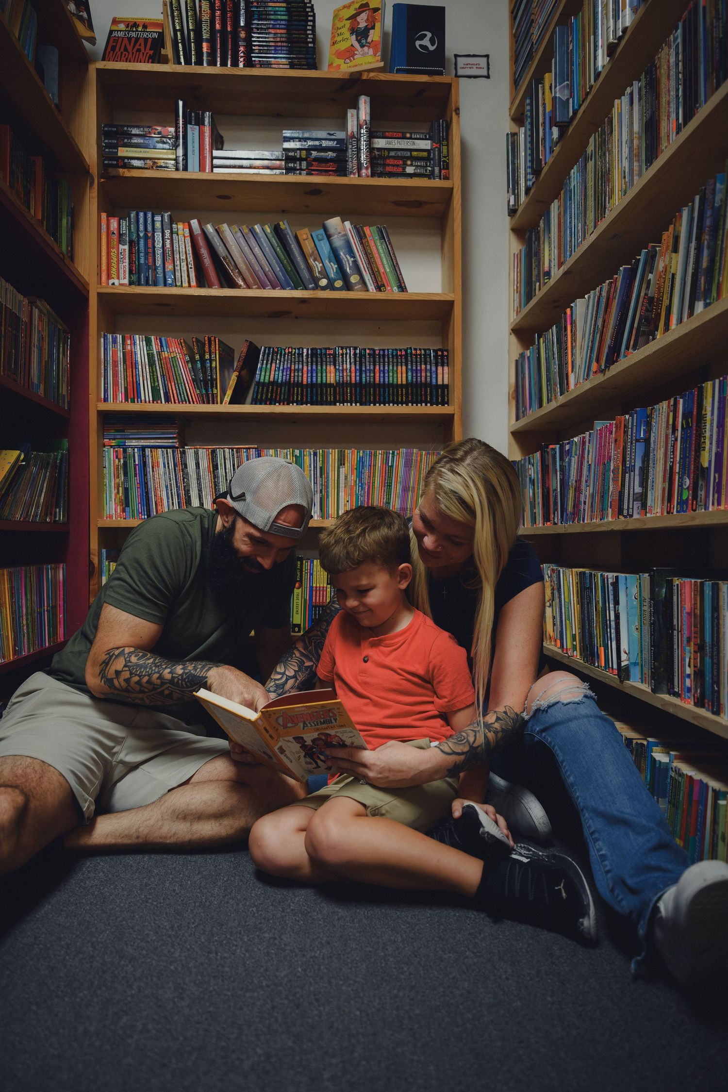 Family reading a book together on the floor of a colorful bookstore library, surrounded by shelves of books.