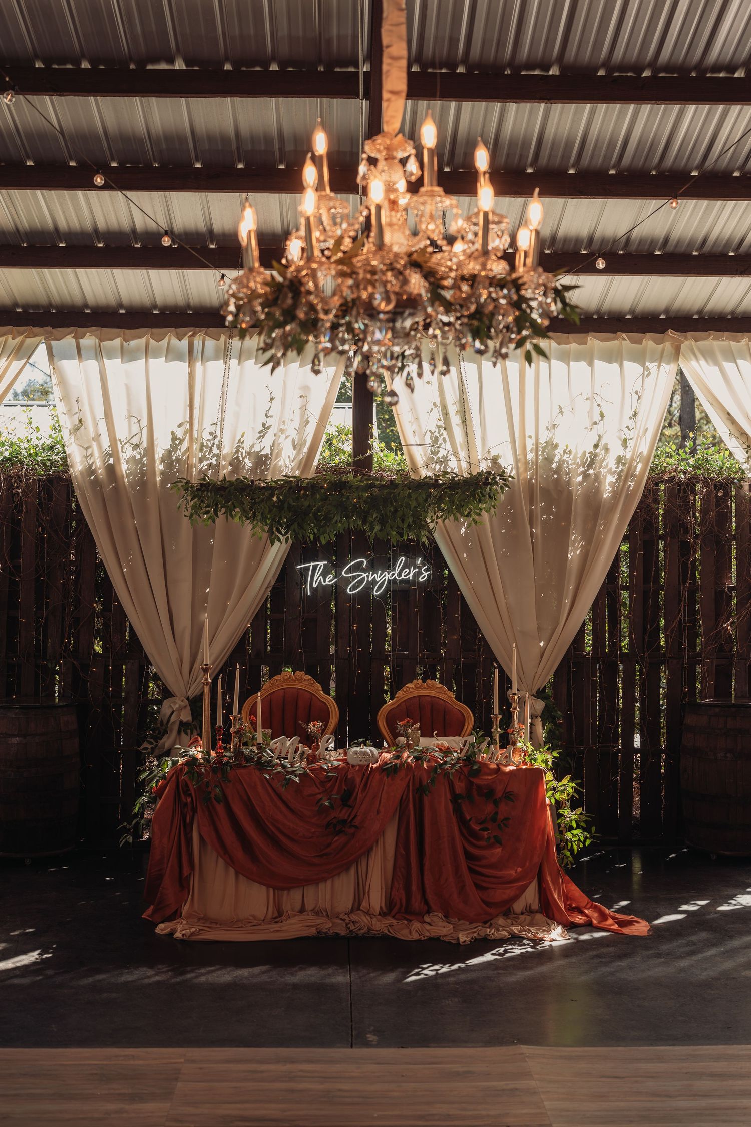Rustic barn wedding sweetheart table with a rust velvet runner, candle chandelier, greenery, ivory drapes, and a neon 