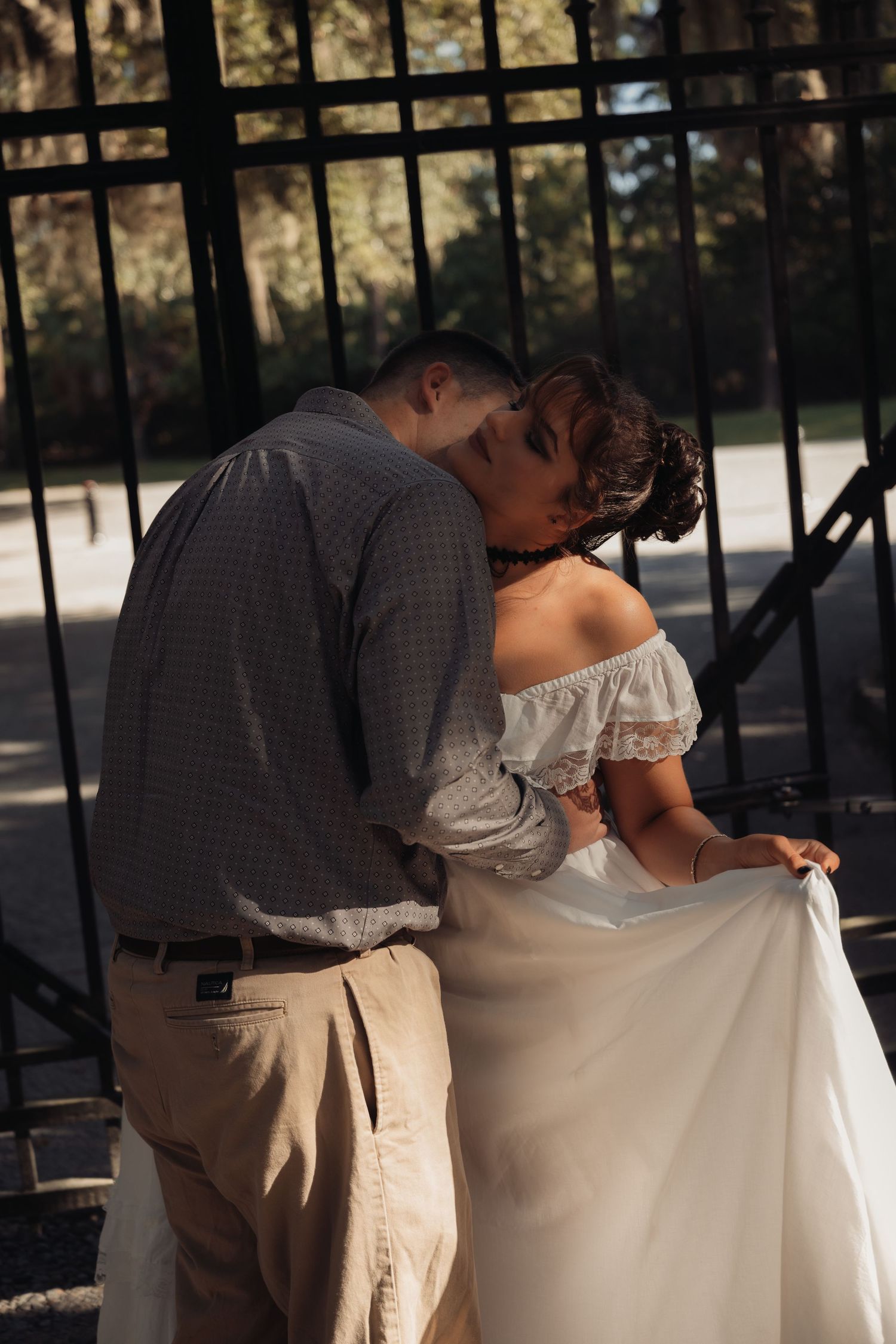 Couple sharing intimate kiss near iron gate in dappled sunlight outdoors.
