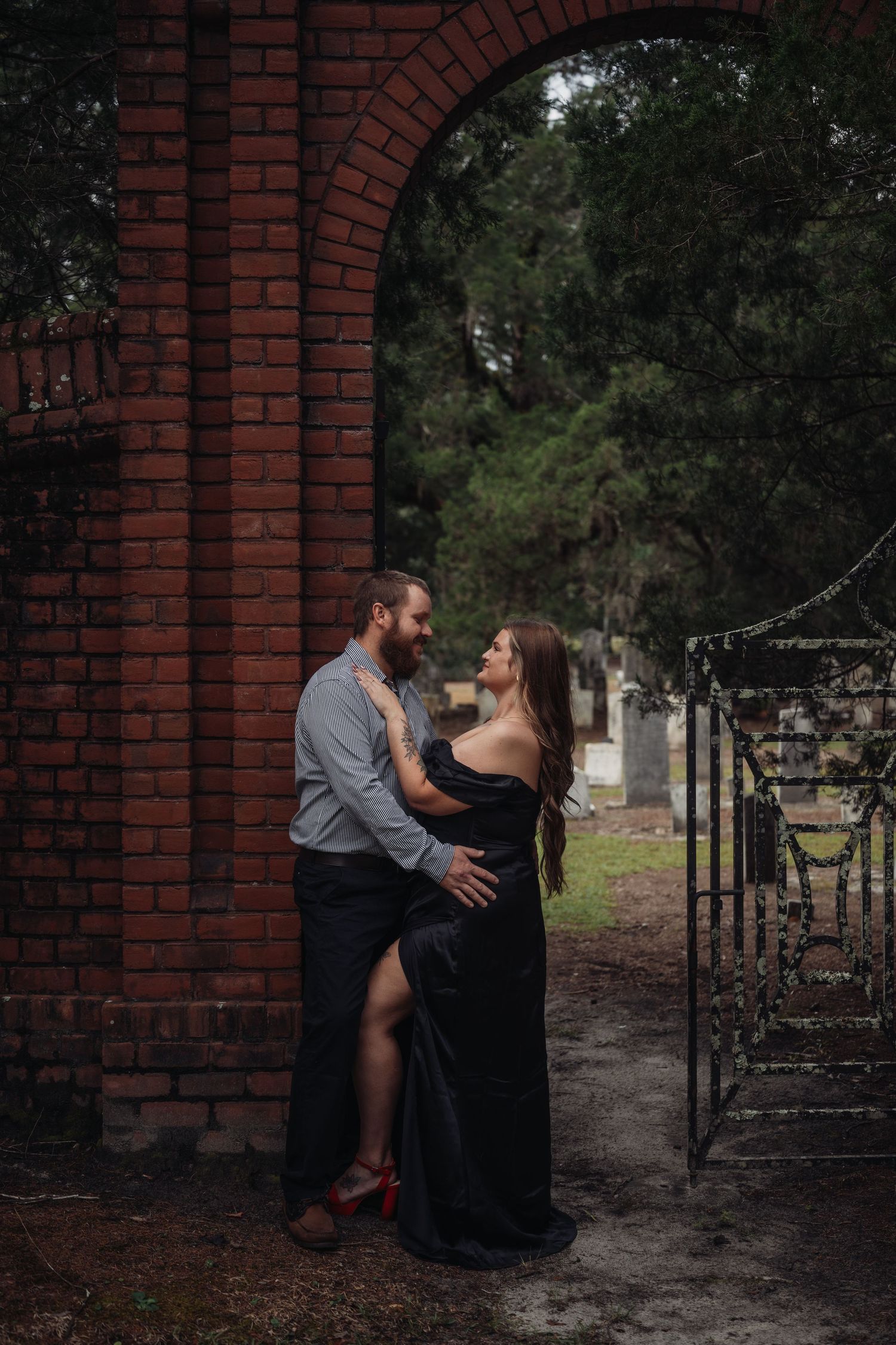 Couple embracing romantically by a brick archway with iron gate, woman in black gown, man in casual attire, outdoor setting.