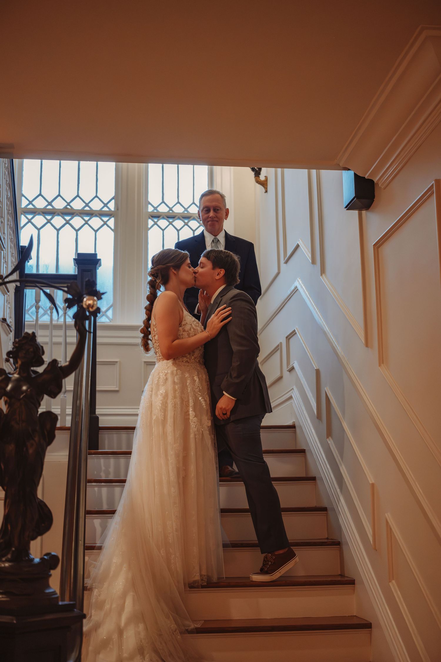 Bride and groom share a kiss on an elegant staircase as an officiant stands behind them during a wedding ceremony.