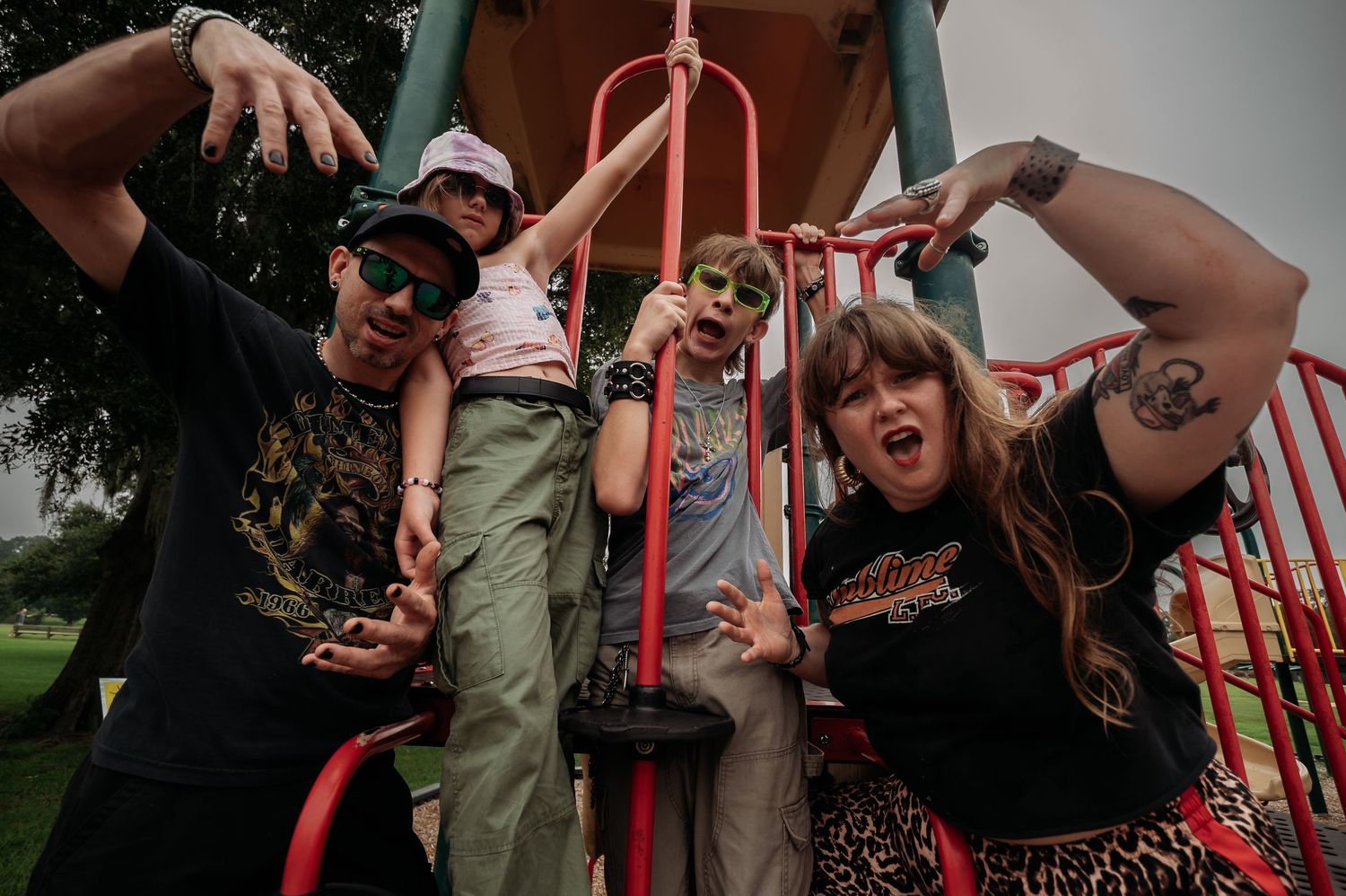 Four people in casual band tees and sunglasses pose playfully on a red playground structure, making expressive faces under a cloudy sky.