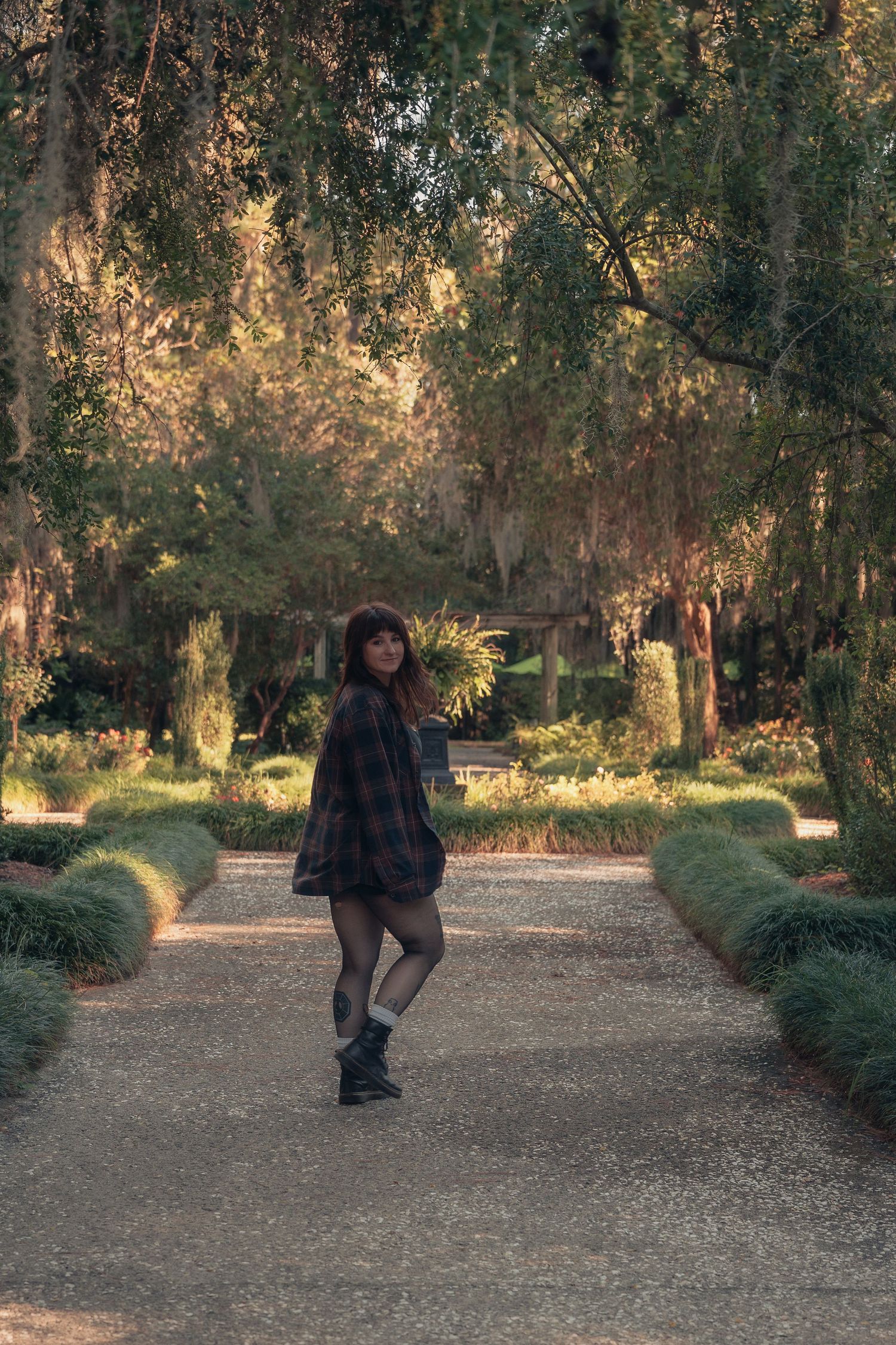 Young woman in plaid shirt and shorts poses on a tree-lined garden path in warm autumn light.