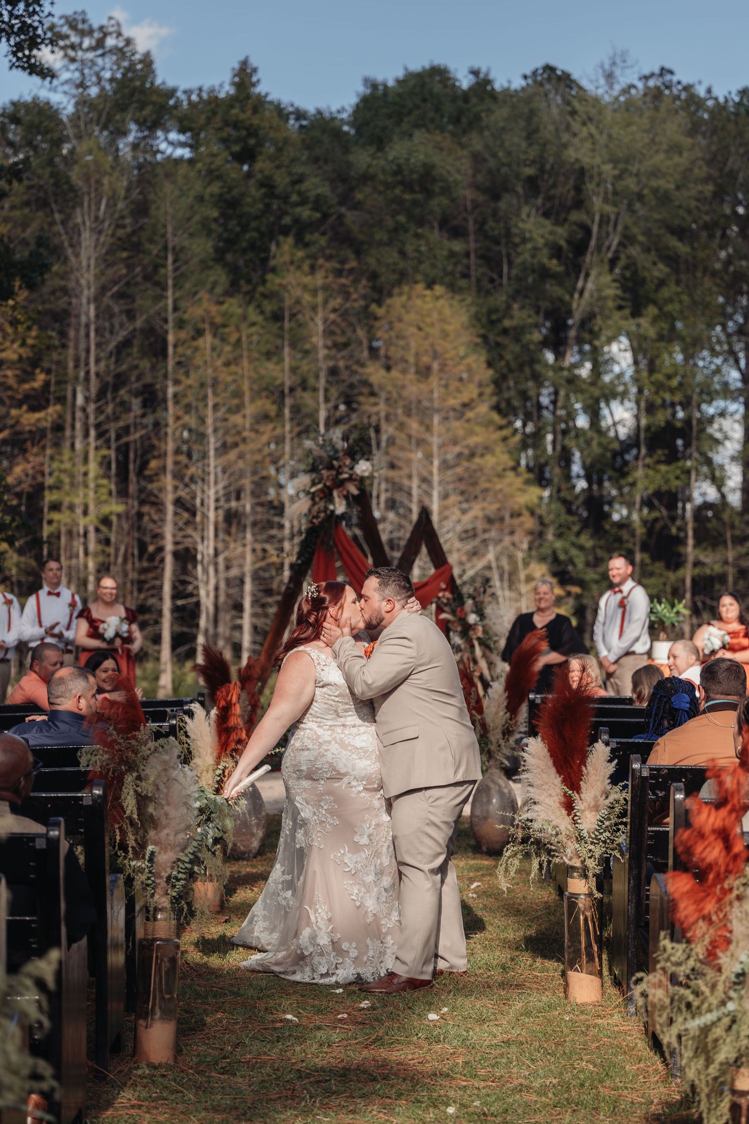 Bride and groom share a kiss in the aisle during an outdoor woodland wedding ceremony as seated guests look on.