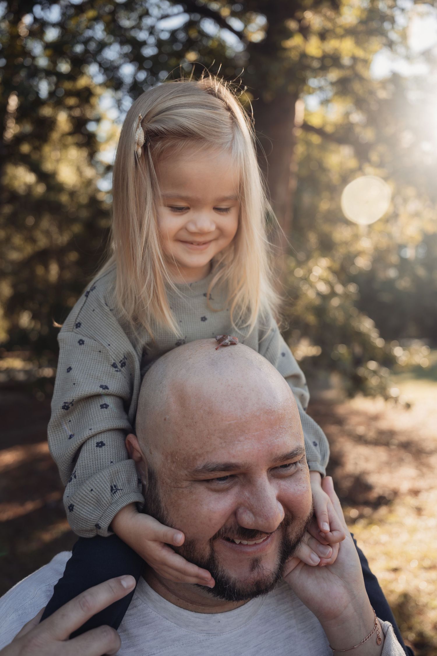 A bald man smiles as his young blonde daughter sits on his shoulders in a sunlit park.