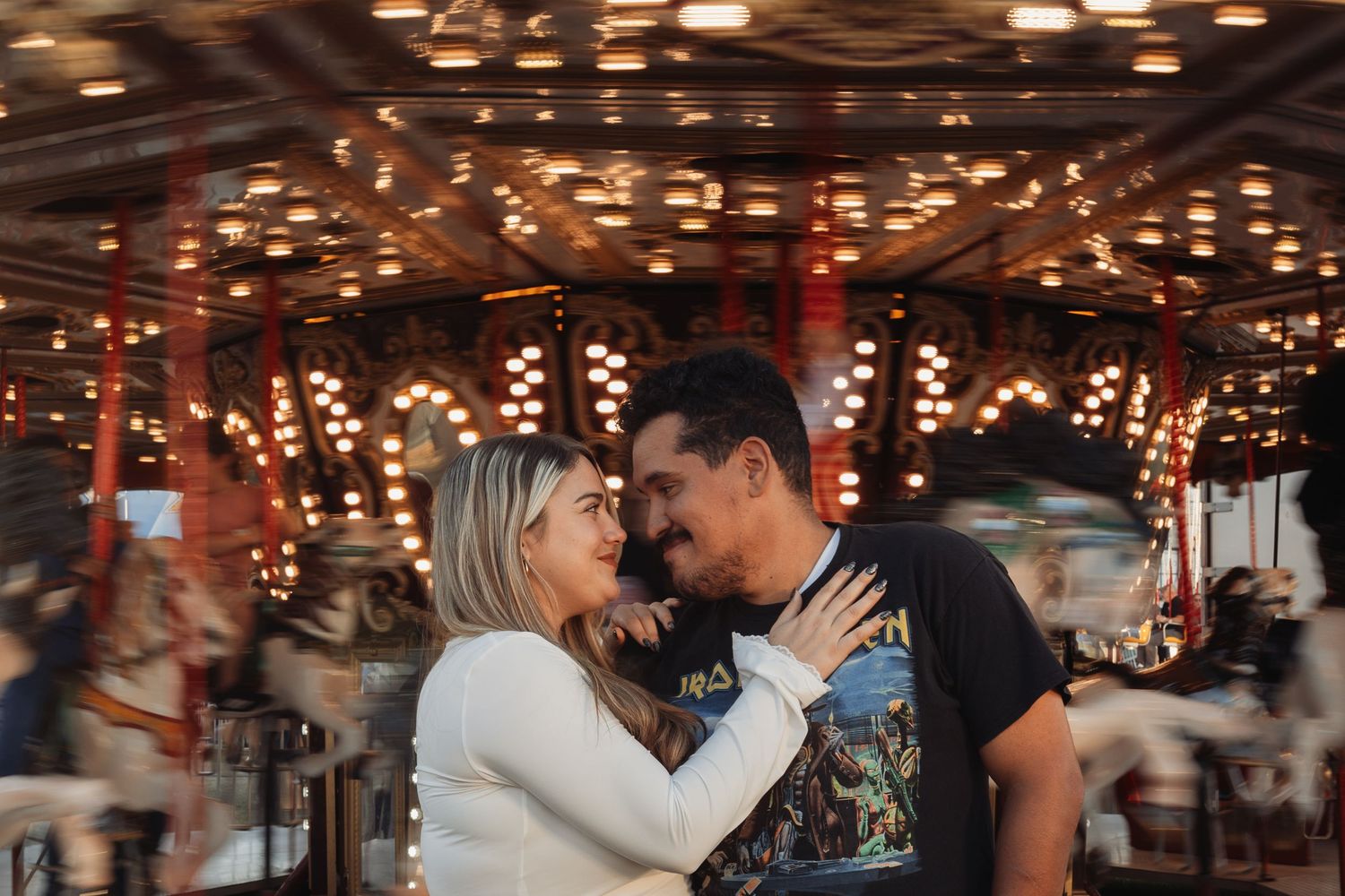 Smiling couple embracing and laughing together in front of a glowing vintage carousel with warm string lights at an amusement park.