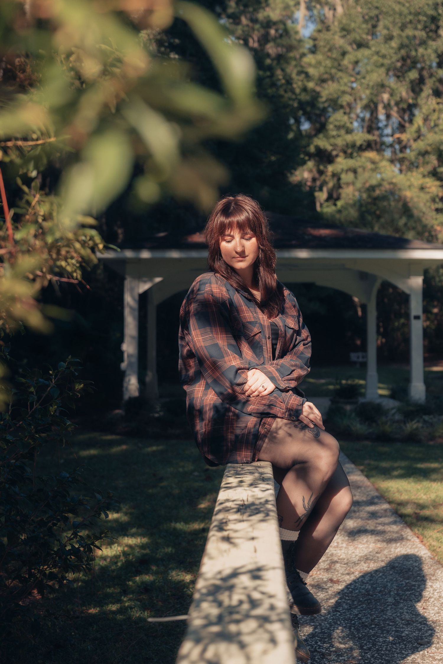 Woman in plaid shirt sits on garden wall with white gazebo and lush green trees in background.