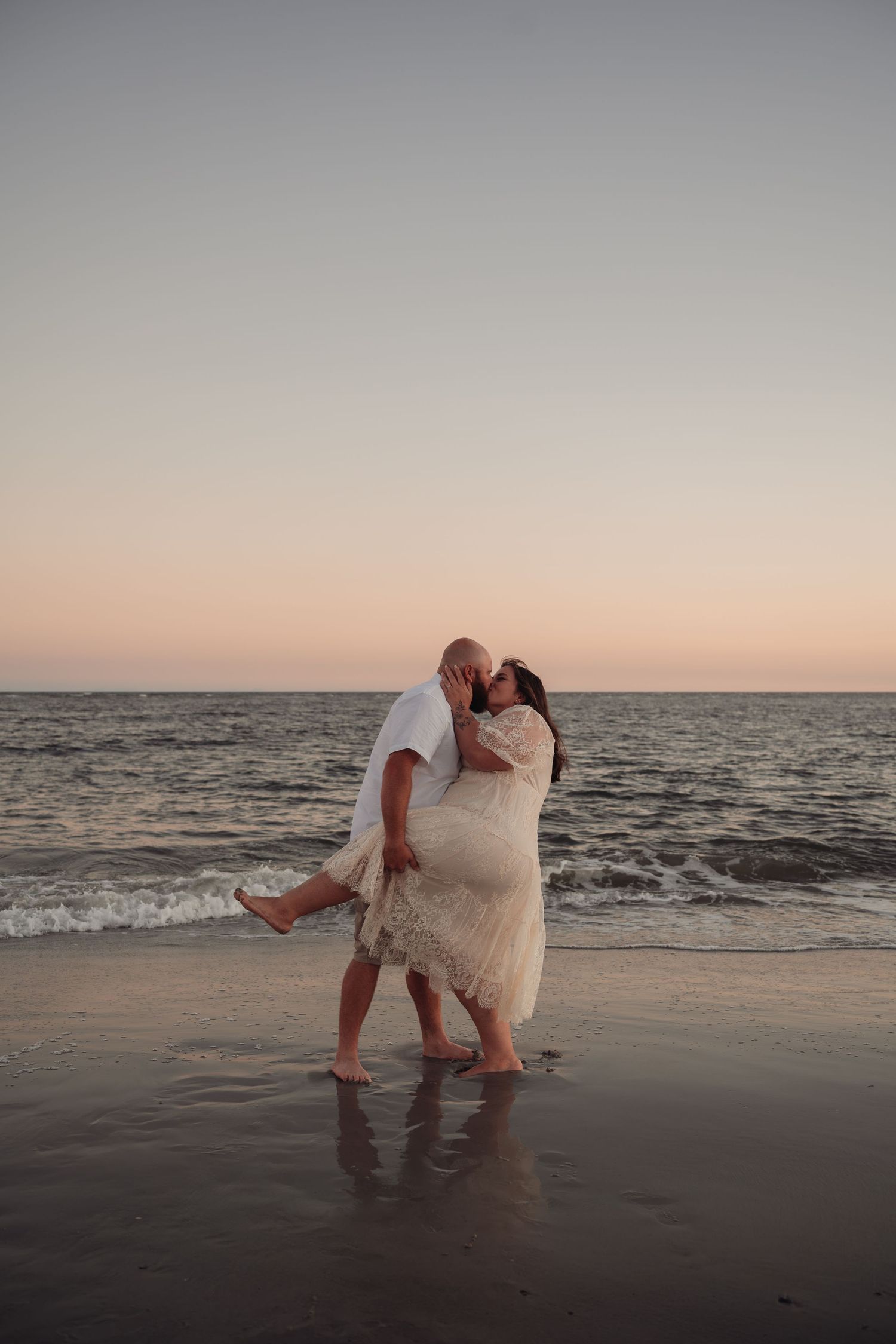 Couple in white outfits kissing on a wet sandy beach at sunset, with gentle waves and a soft pink sky behind them.