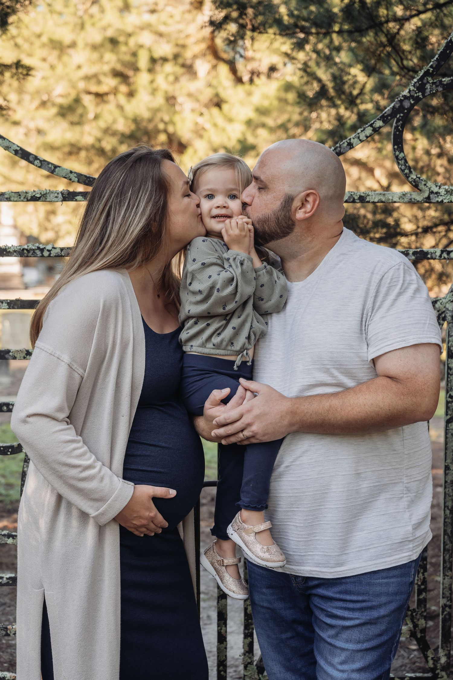Pregnant woman and bald man kissing toddler on cheeks outdoors near iron fence with trees in background.