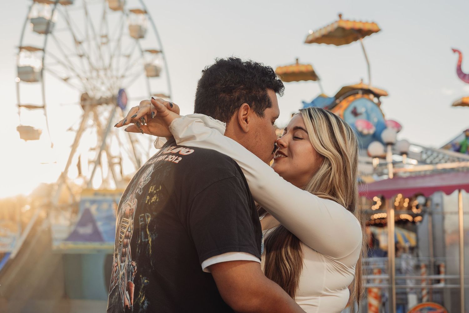 Couple embracing and kissing at a sunset carnival with a Ferris wheel and festive amusement park rides glowing in the background.
