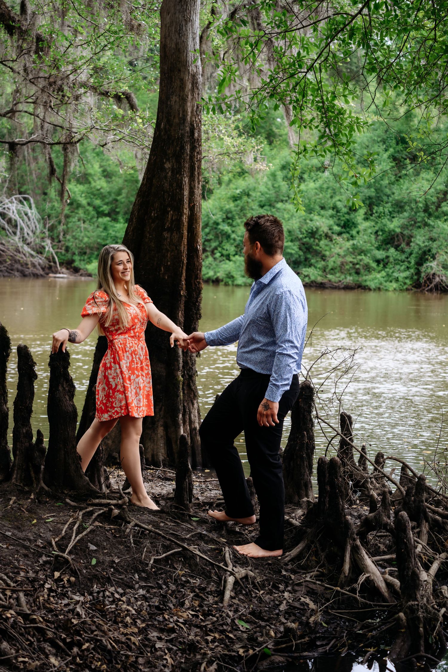 Couple holds hands barefoot on cypress tree roots beside a serene river surrounded by green trees.