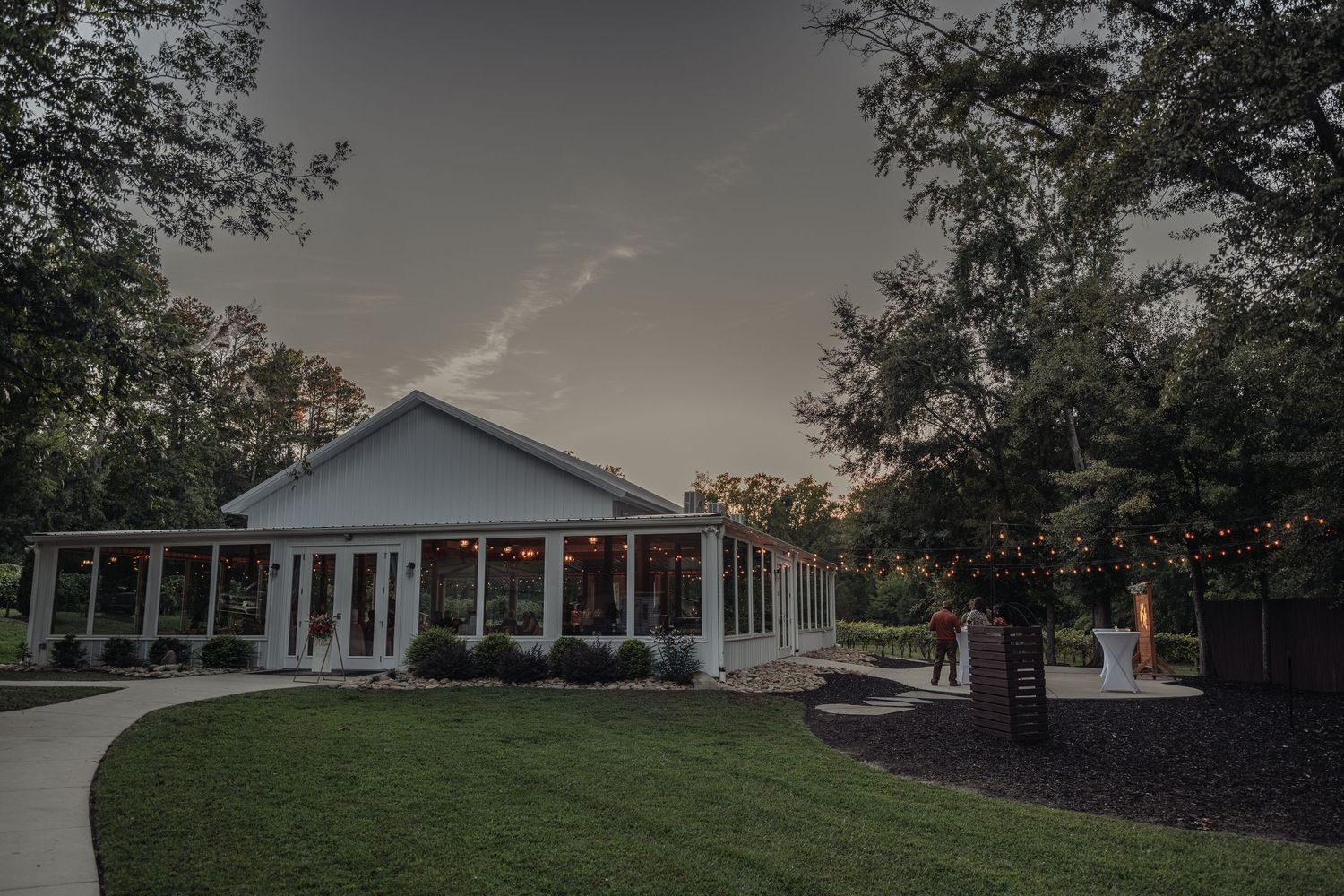 Elegant white wedding venue with string lights glowing at dusk, surrounded by lush trees.