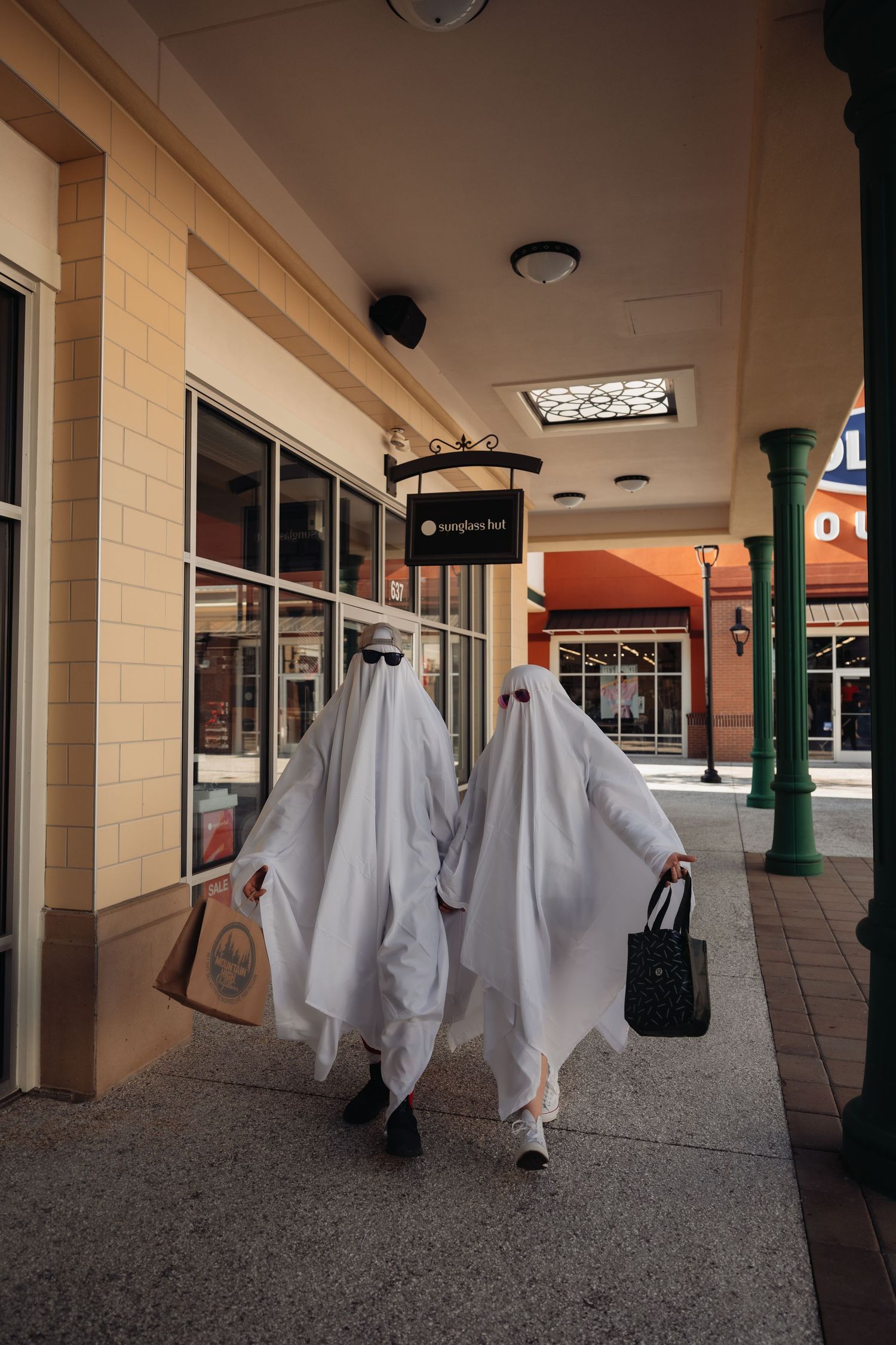 Two people in white ghost costumes walk through an outdoor shopping mall carrying bags on Halloween.