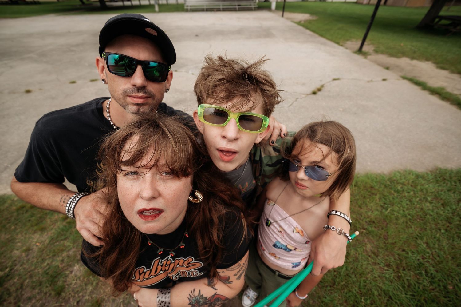 Four friends in sunglasses huddle close for a fisheye-style group photo outdoors on a paved lot with green grass in the background.