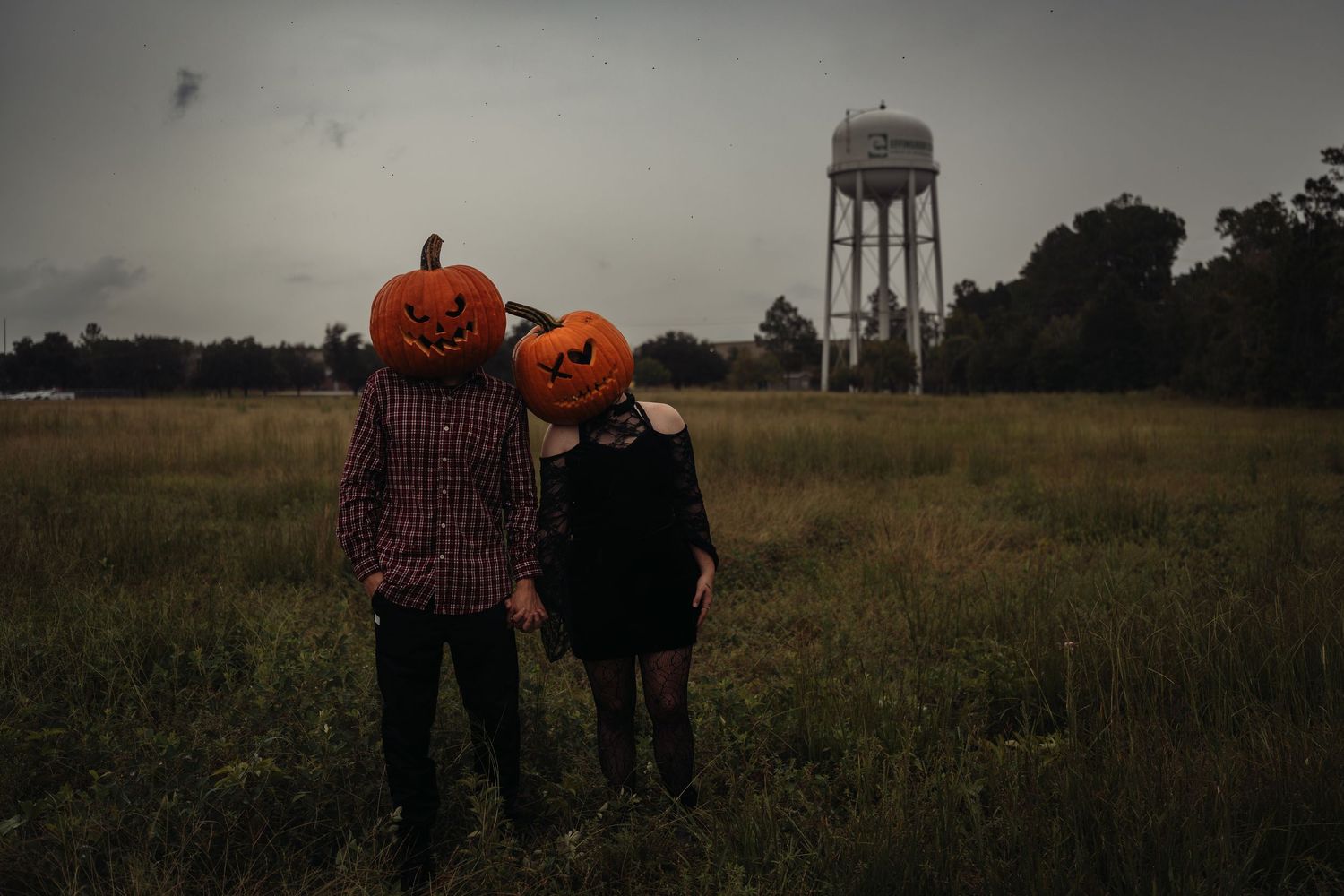 Couple wearing carved Halloween pumpkins on heads standing in a grassy field with a water tower in the background.