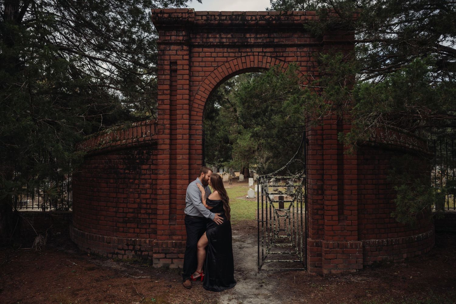 Couple sharing a romantic kiss under a rustic brick archway with iron gate during an outdoor maternity photoshoot.