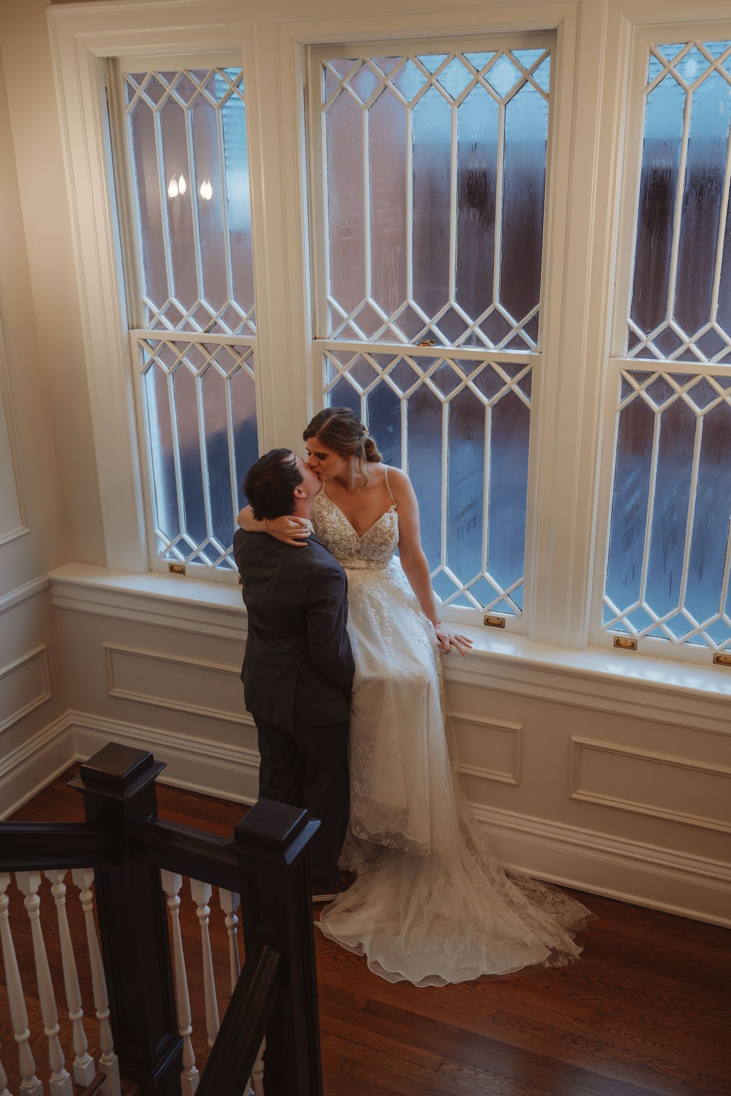 Bride and groom share a romantic moment by elegant leaded glass windows on a staircase landing in a classic venue.