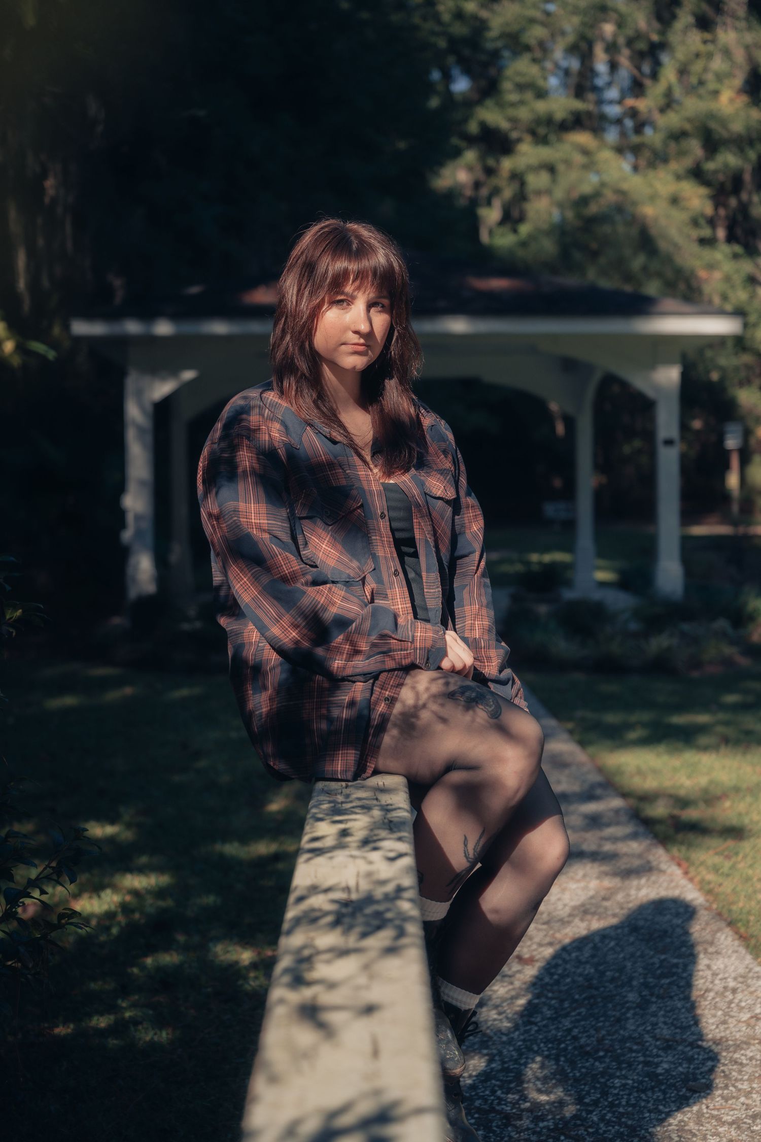 Young woman in plaid shirt sits on stone ledge in sunlit garden with white gazebo backdrop.