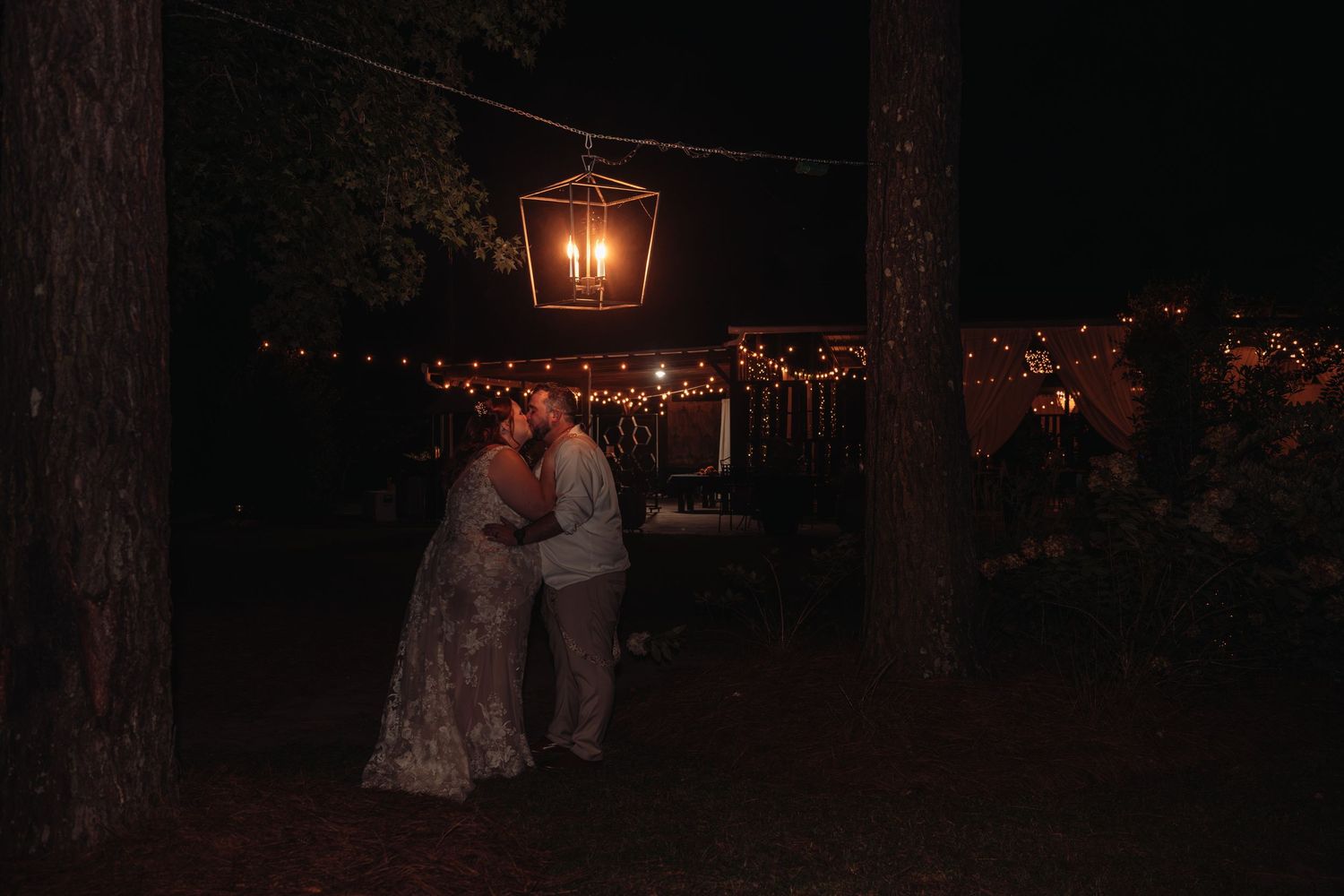 Couple in wedding attire embrace under a hanging lantern and string lights at a dark outdoor evening reception among tall trees.
