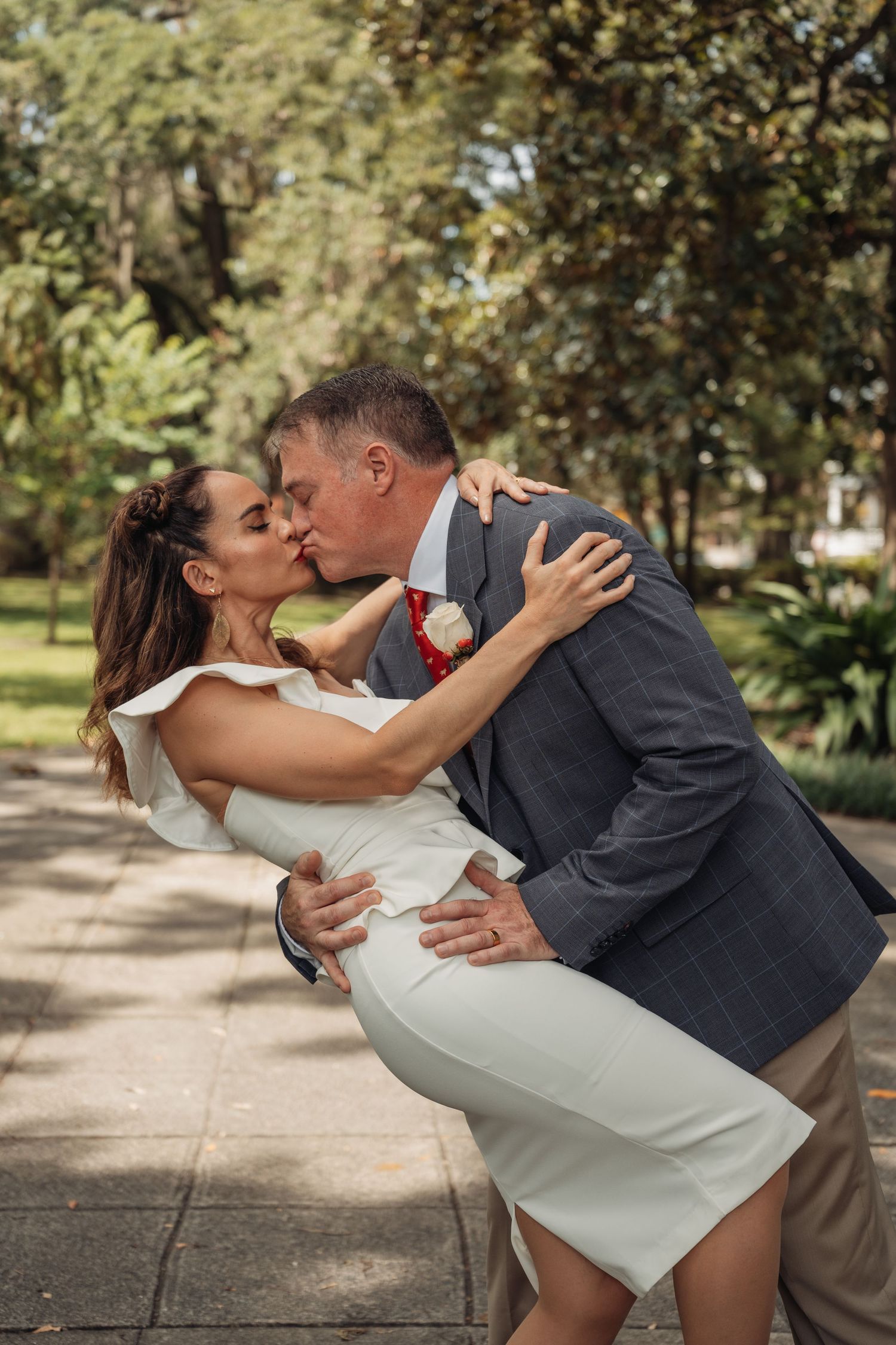 A groom dips his bride for a romantic kiss along a sunlit park pathway surrounded by lush green trees.