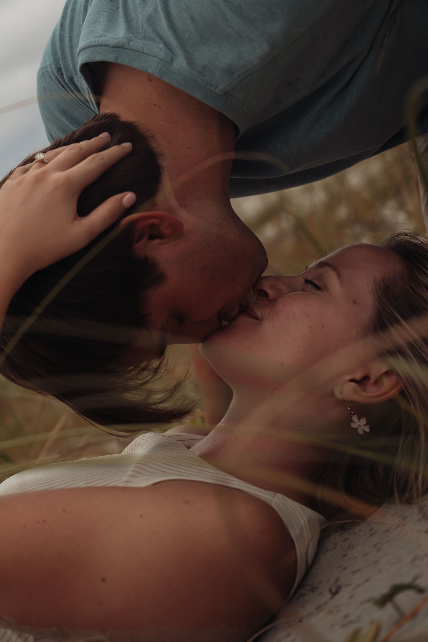 A couple shares a tender kiss outdoors, with the woman lying back and the man leaning in, surrounded by natural scenery.