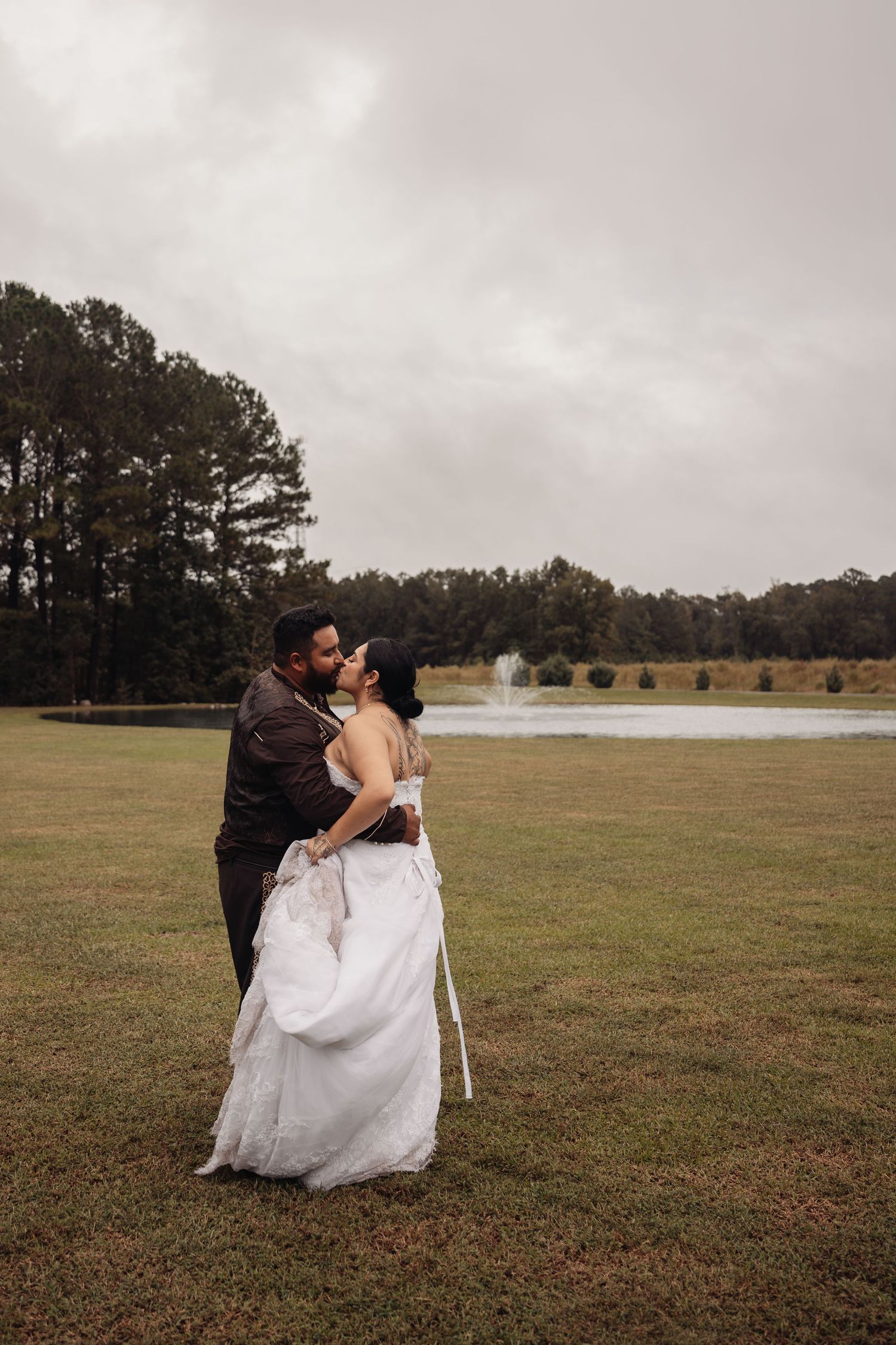 Bride and groom share a romantic kiss outdoors on a grassy lawn with a pond and fountain backdrop on a cloudy day.