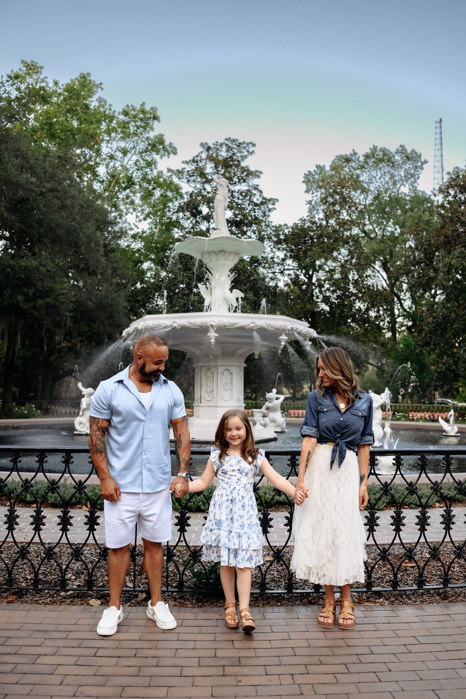 A family of three poses hand-in-hand before a white fountain in a lush park, dressed in coordinating blue and white outfits.