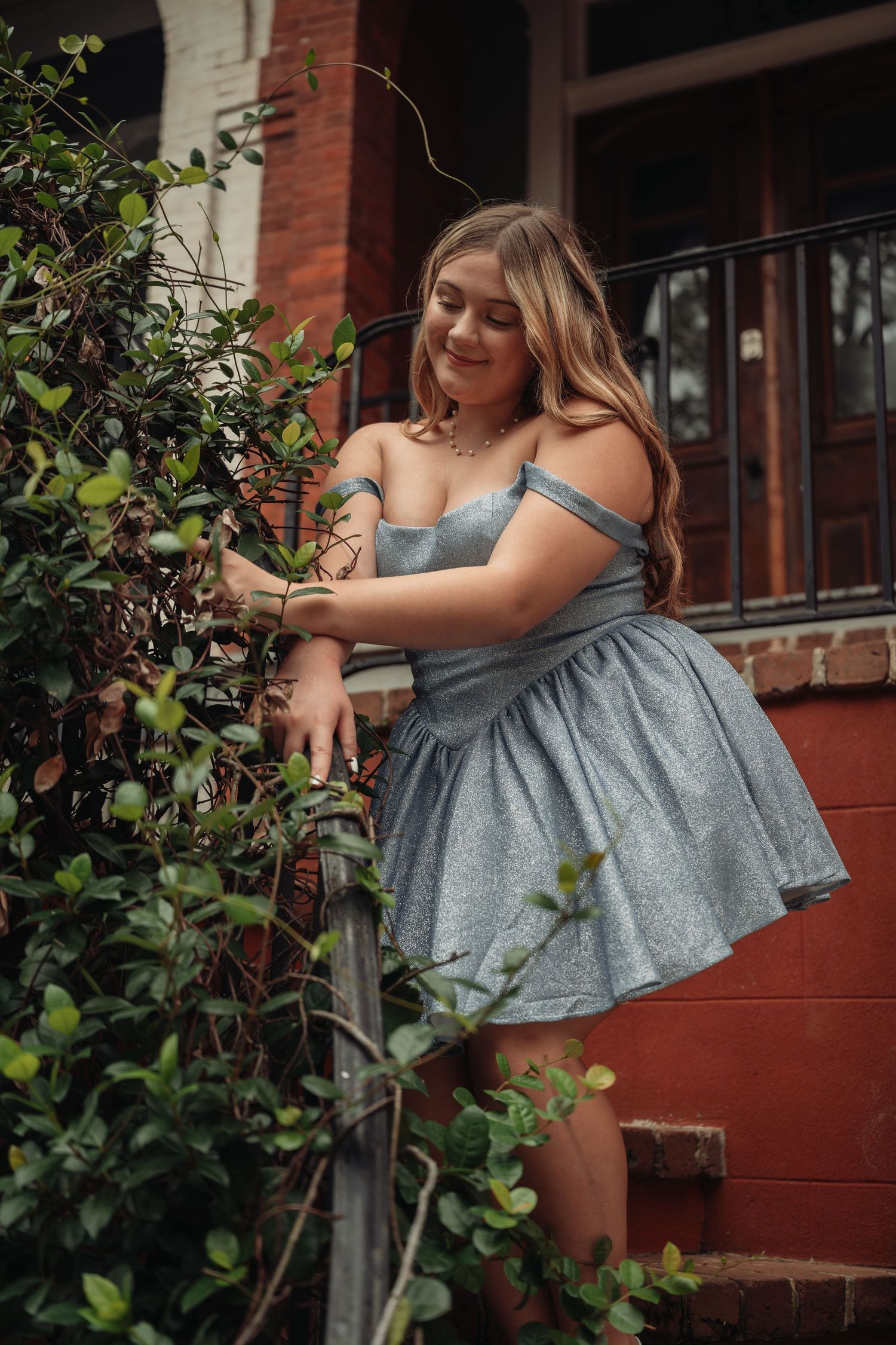 Young woman in a silver off-shoulder mini dress smiling among green bushes near a red brick building.