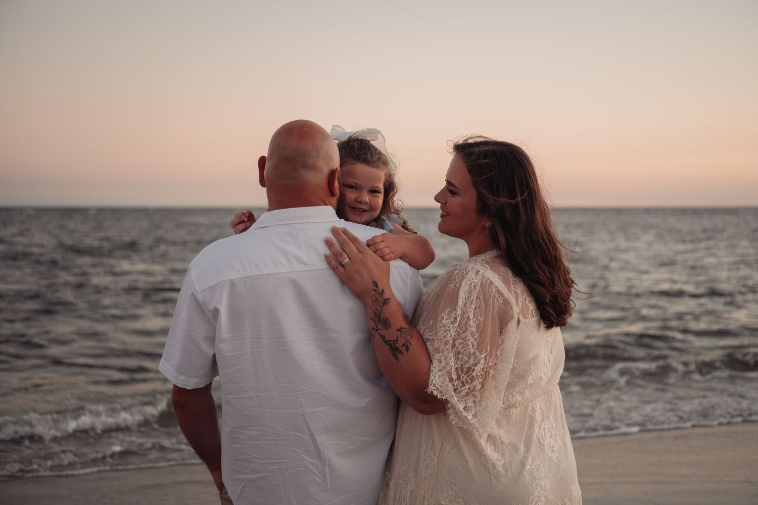 Family of three embracing on a beach at sunset, with a smiling woman in white lace holding a young girl beside a man in a white shirt.