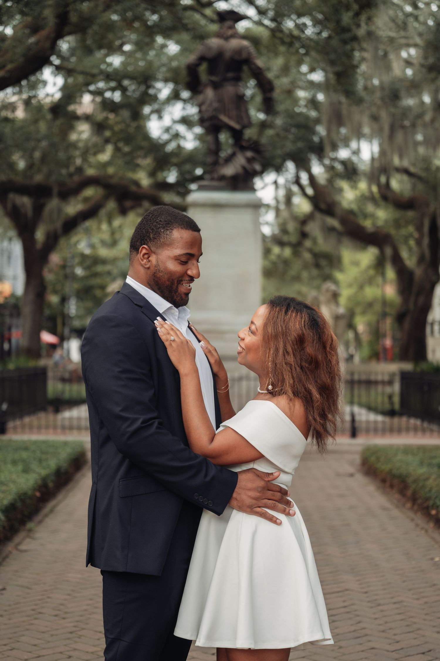 Couple embracing romantically in Savannah park with statue backdrop, man in navy suit, woman in white off-shoulder dress.