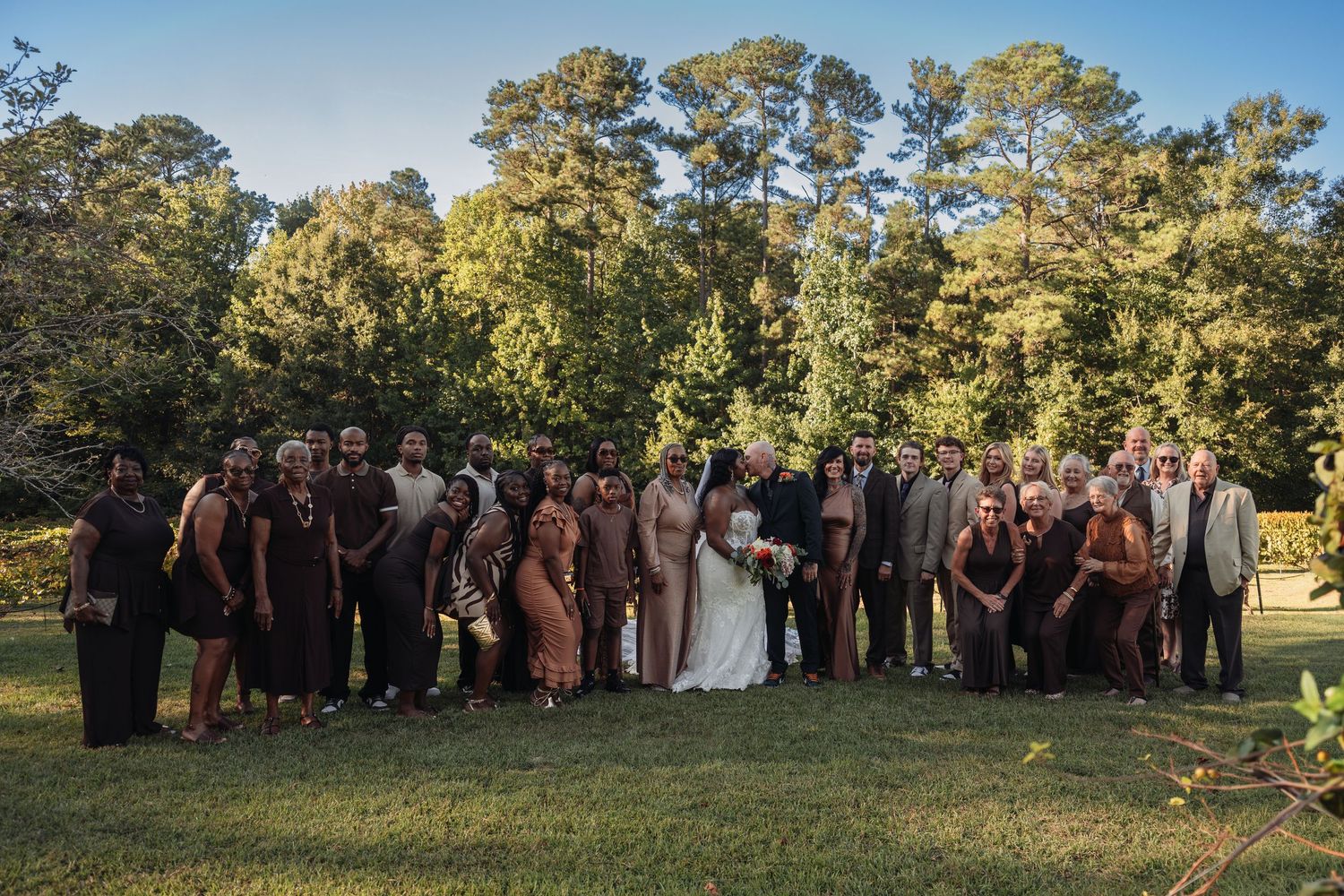 Large interracial wedding party group photo on green lawn surrounded by tall trees.