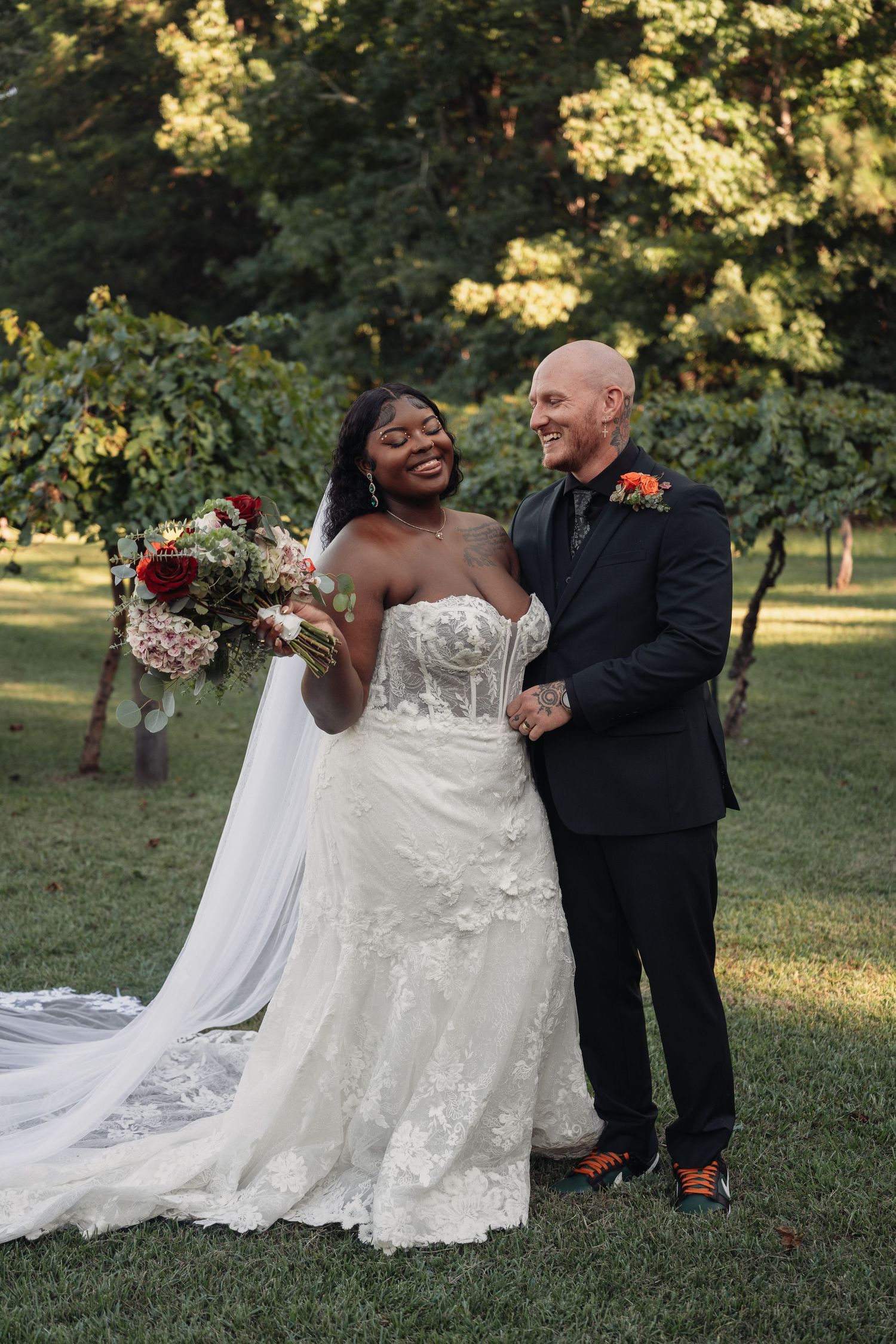 Smiling interracial couple poses together in a garden, bride holding a red and white bouquet.