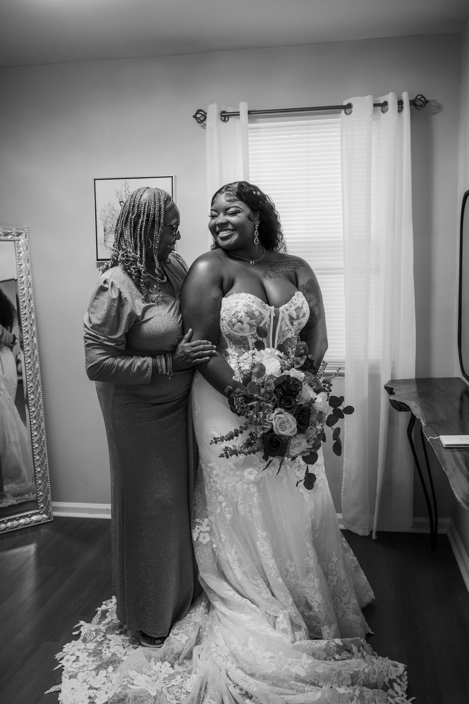 Black and white photo of a smiling bride in lace gown holding bouquet with a woman beside her.