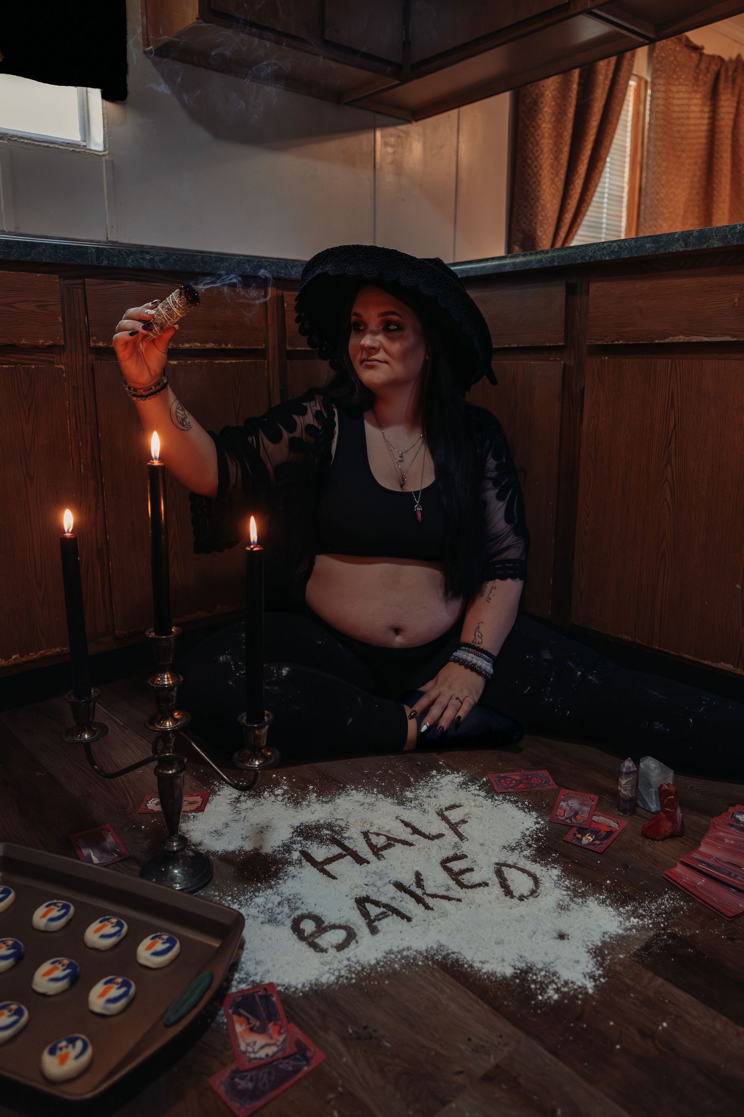Woman in witch hat poses on floor with candles and flour spelling Half Baked in dark moody kitchen.