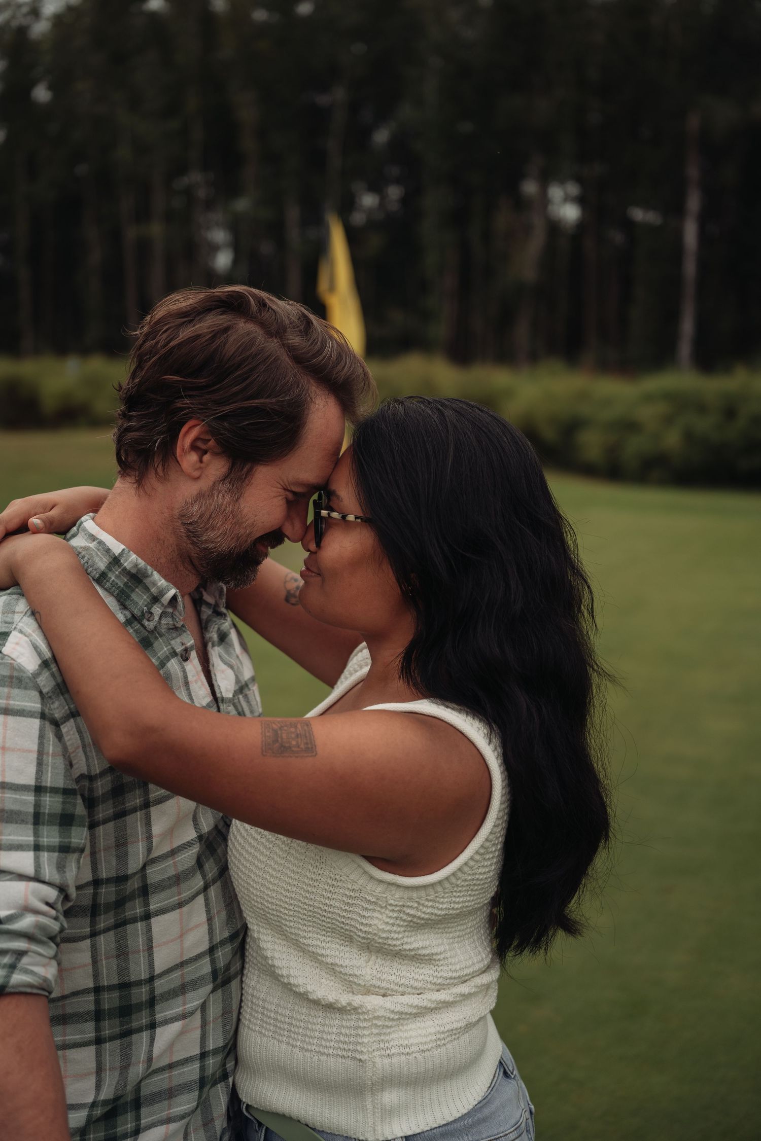 A couple shares a romantic forehead touch on a lush golf course, with tall trees in the background.