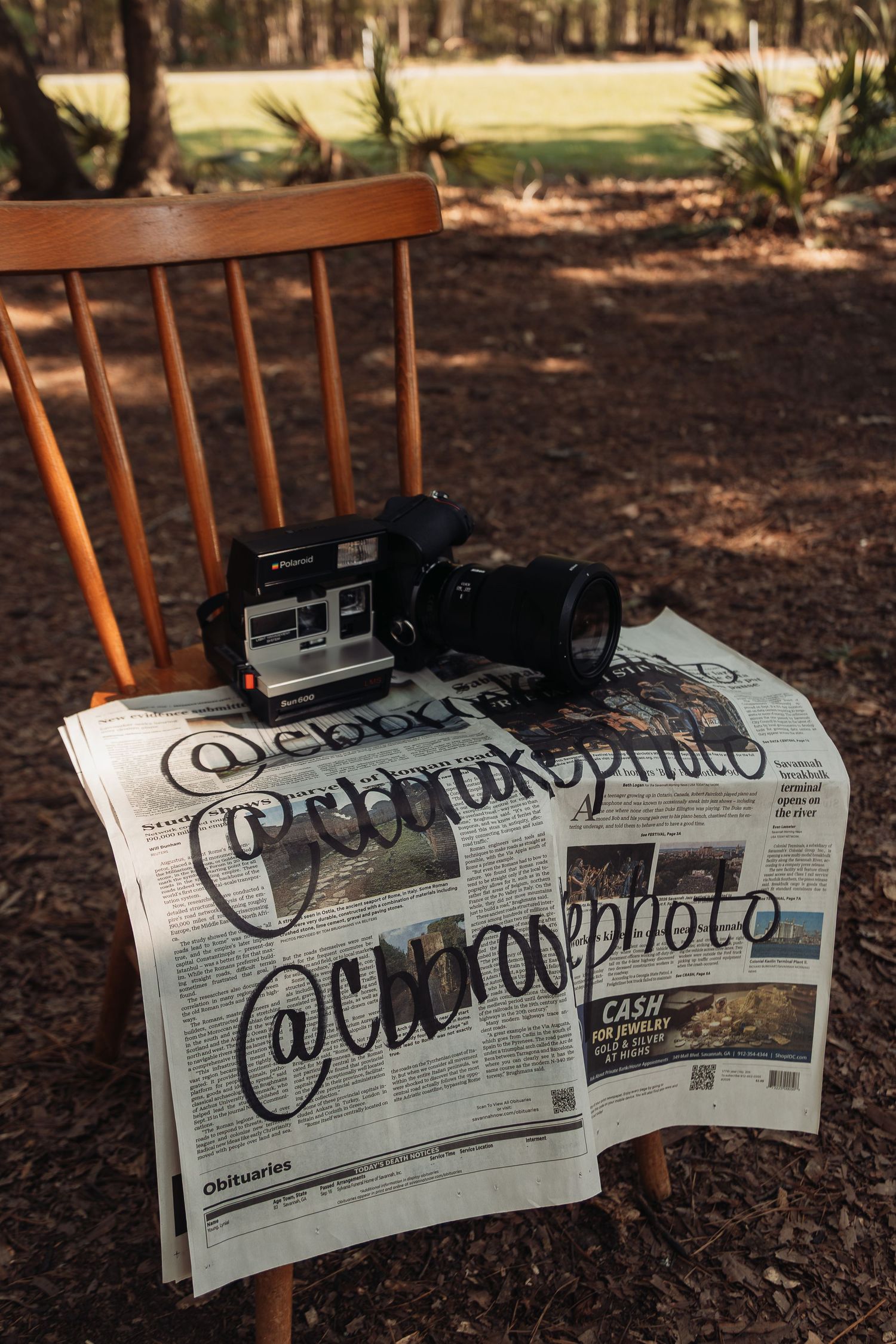Vintage cameras and newspaper with social media handle @cbrosephoto displayed on wooden chair in forest.