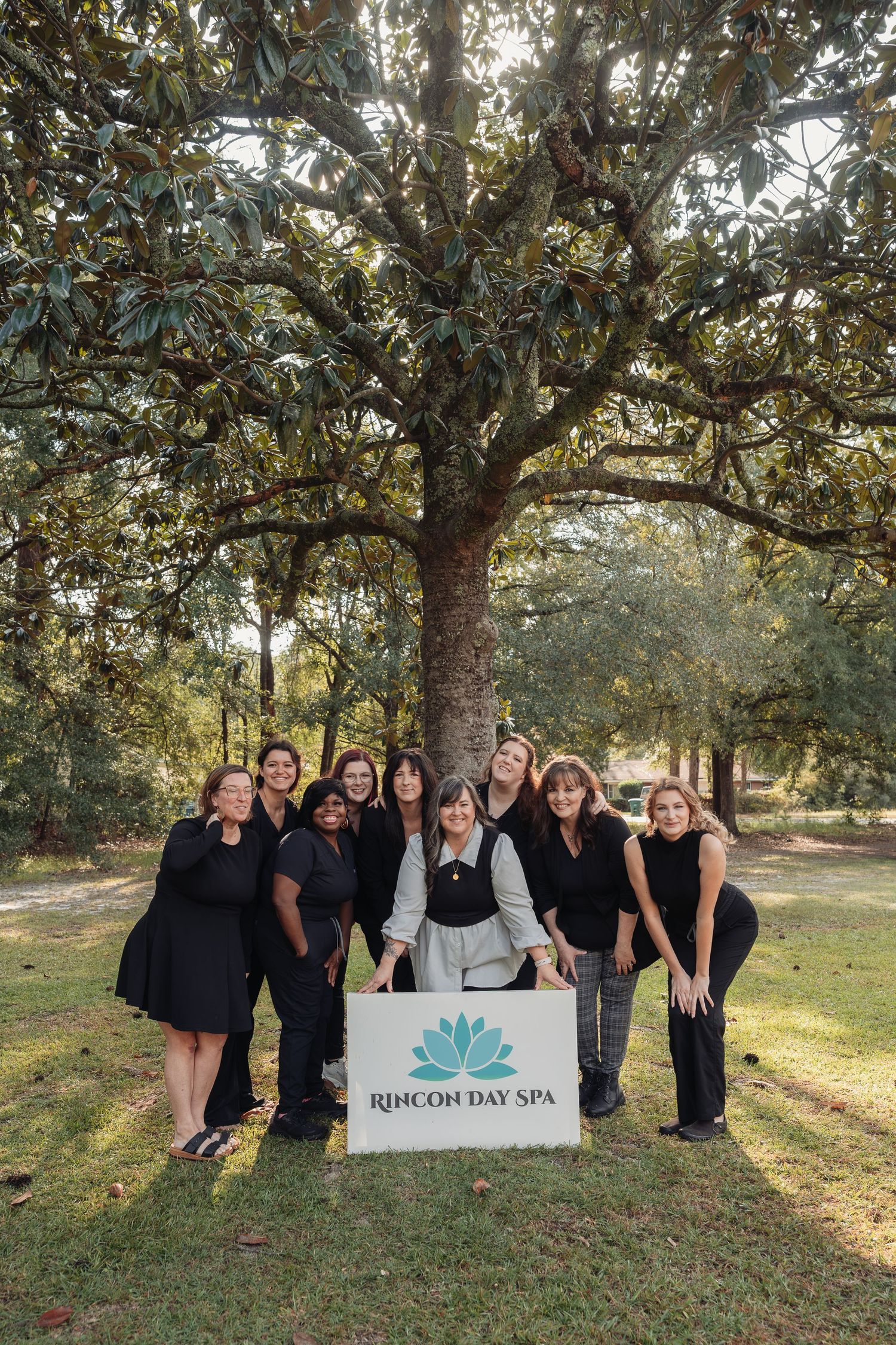 Rincon Day Spa team members in black uniforms pose together under a large magnolia tree holding a spa logo sign.