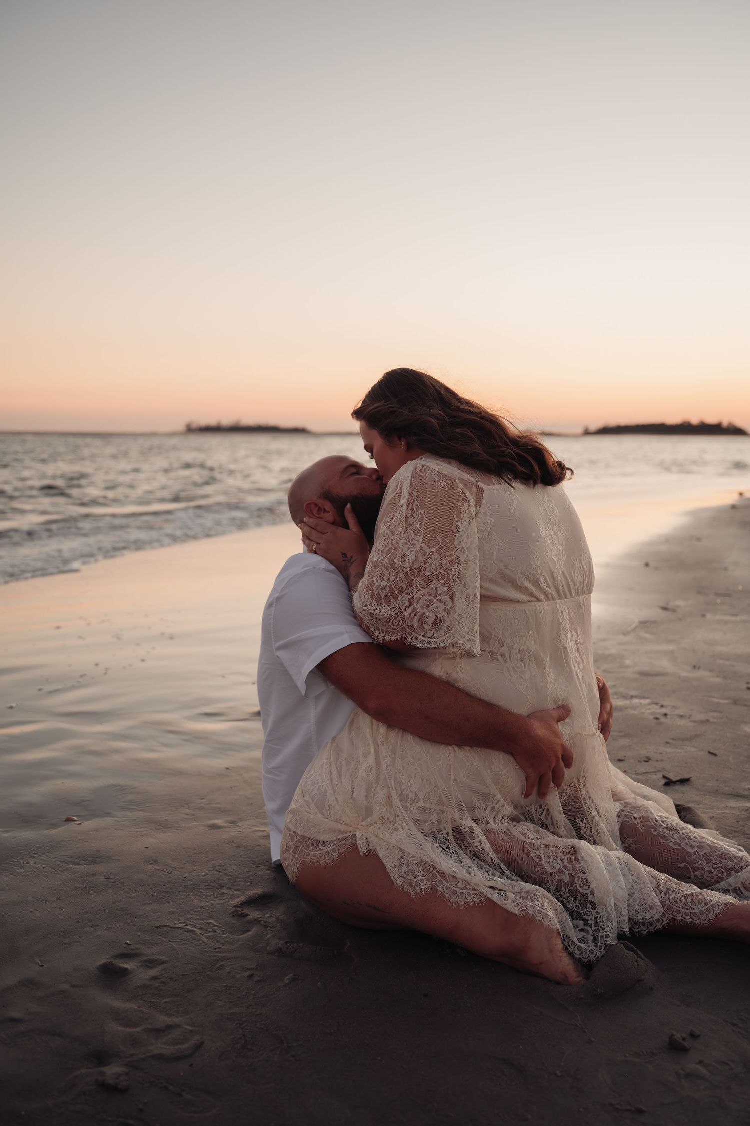 Couple in white embrace and kiss while sitting on a sandy beach at sunset, with calm ocean waves and distant islands behind them.