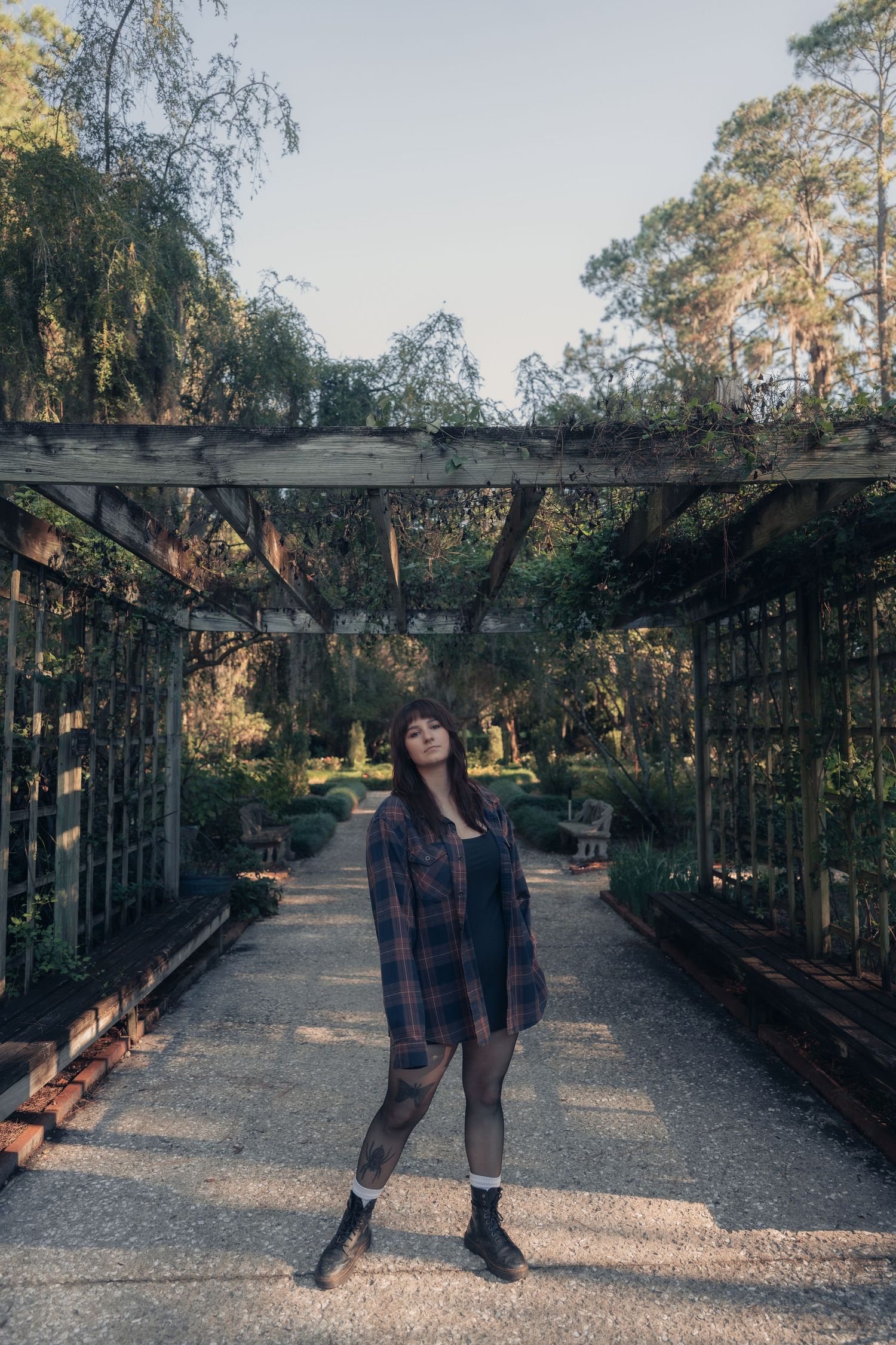 Young woman in plaid shirt and black boots posing under a rustic garden pergola surrounded by lush greenery.